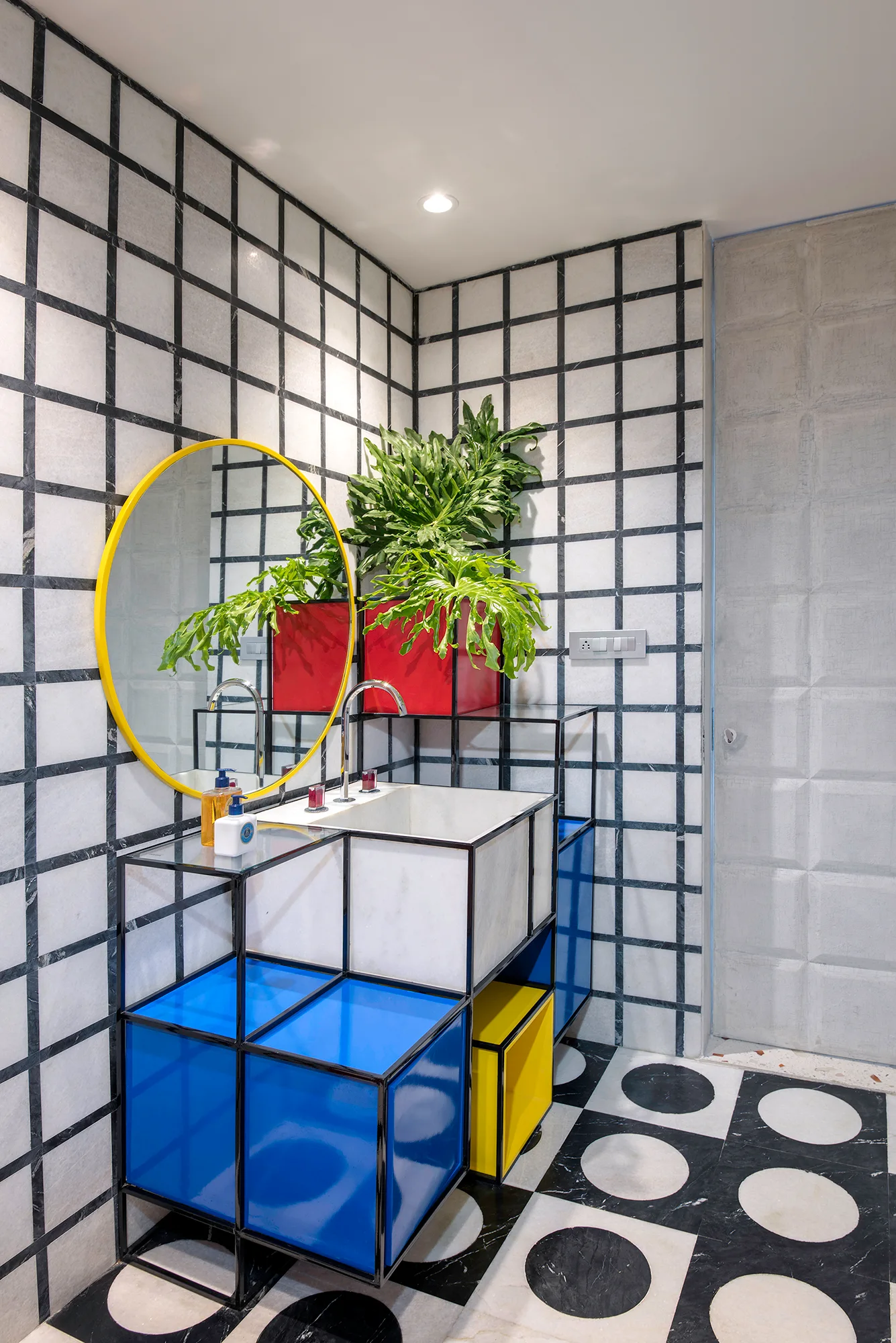 A powder room features a modular black metal vanity with blue and yellow panels, a round yellow-rimmed mirror, and grid-pattern marble wall tiles.