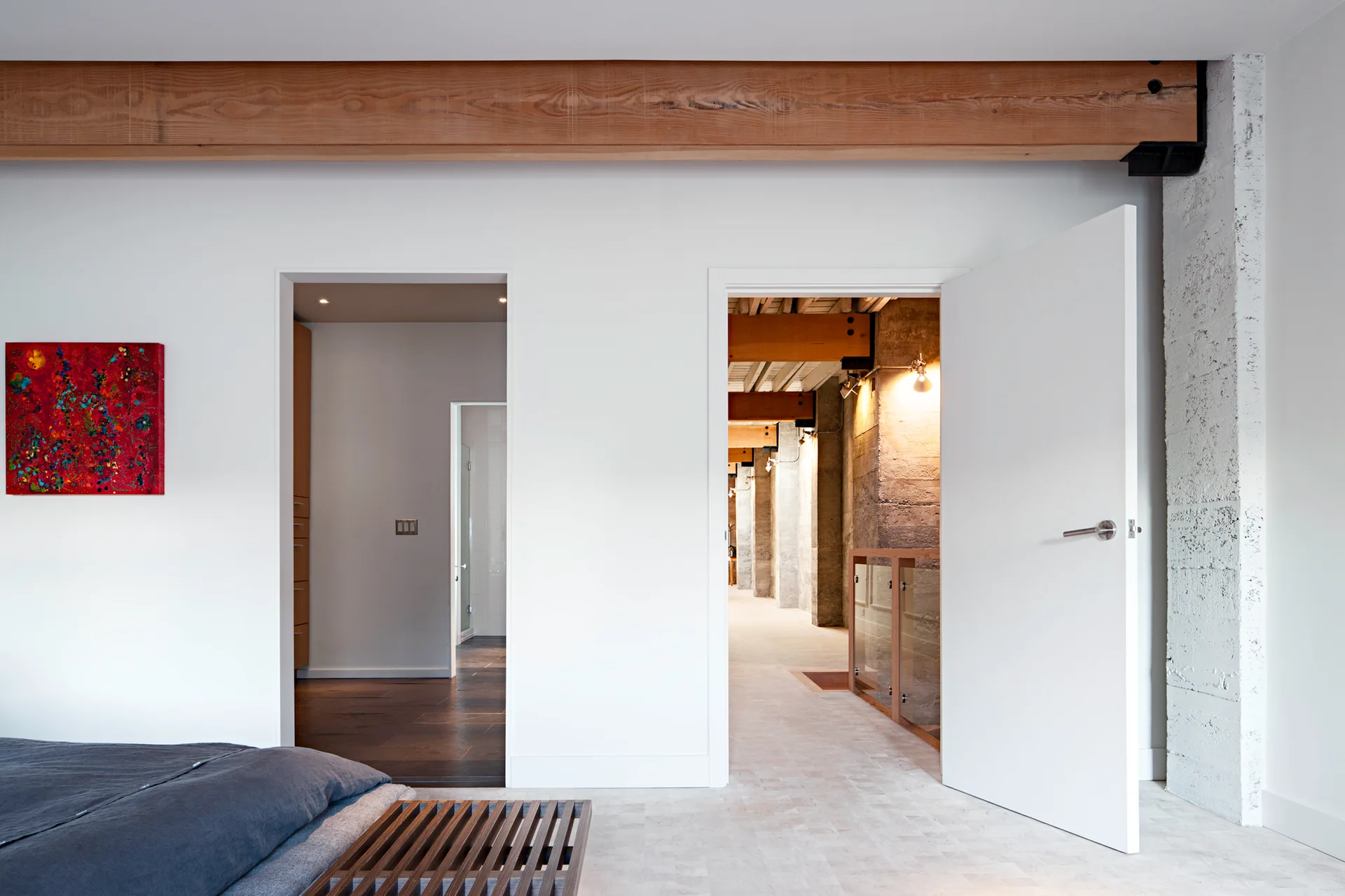 A minimalist bedroom features white walls, a large exposed wooden ceiling beam, a textured concrete pillar, and an open doorway leading to a hallway.