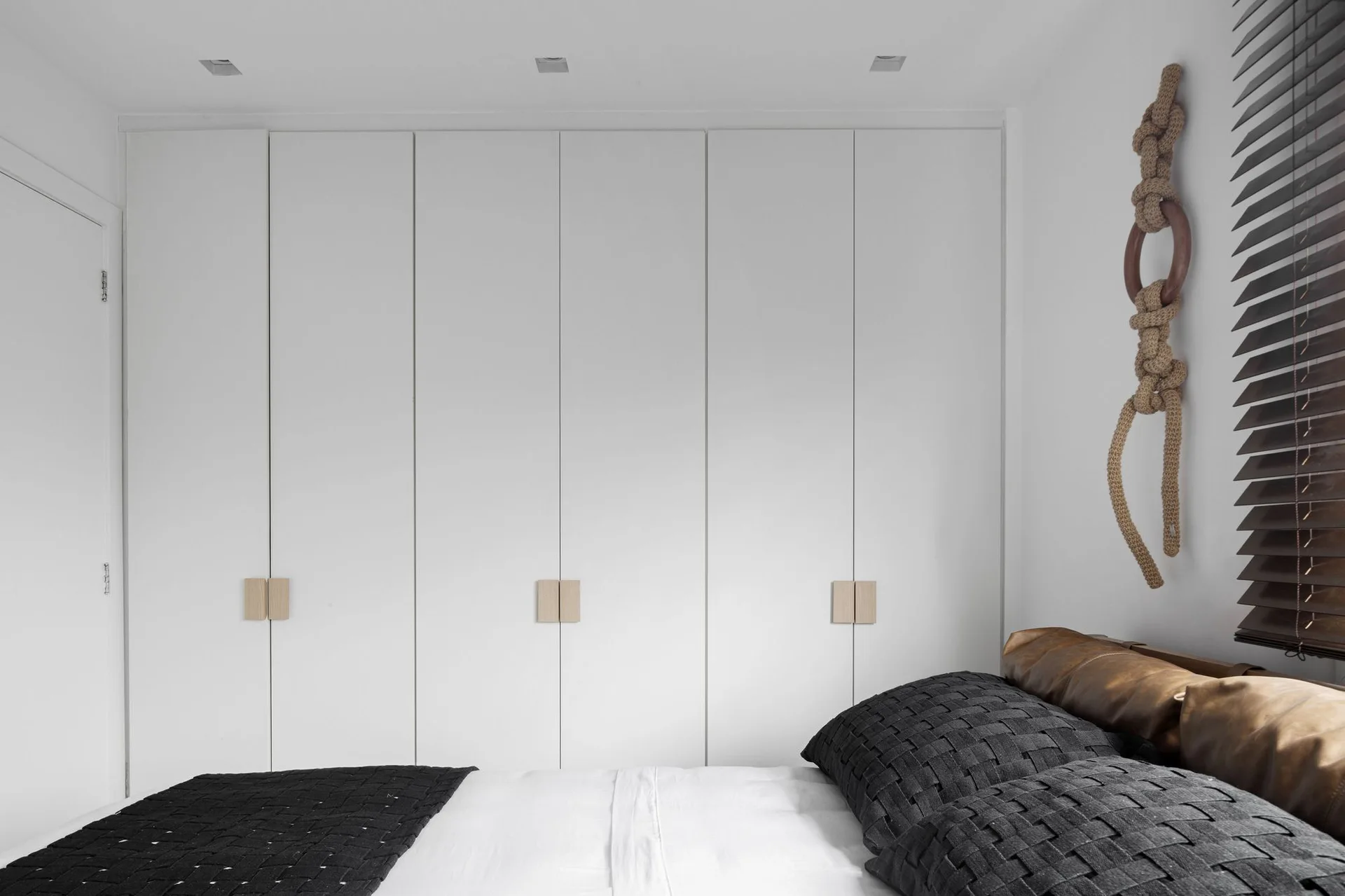 A minimalist bedroom features a wall of floor-to-ceiling white wardrobes with wood square pulls alongside a bed styled with black and brown pillows.