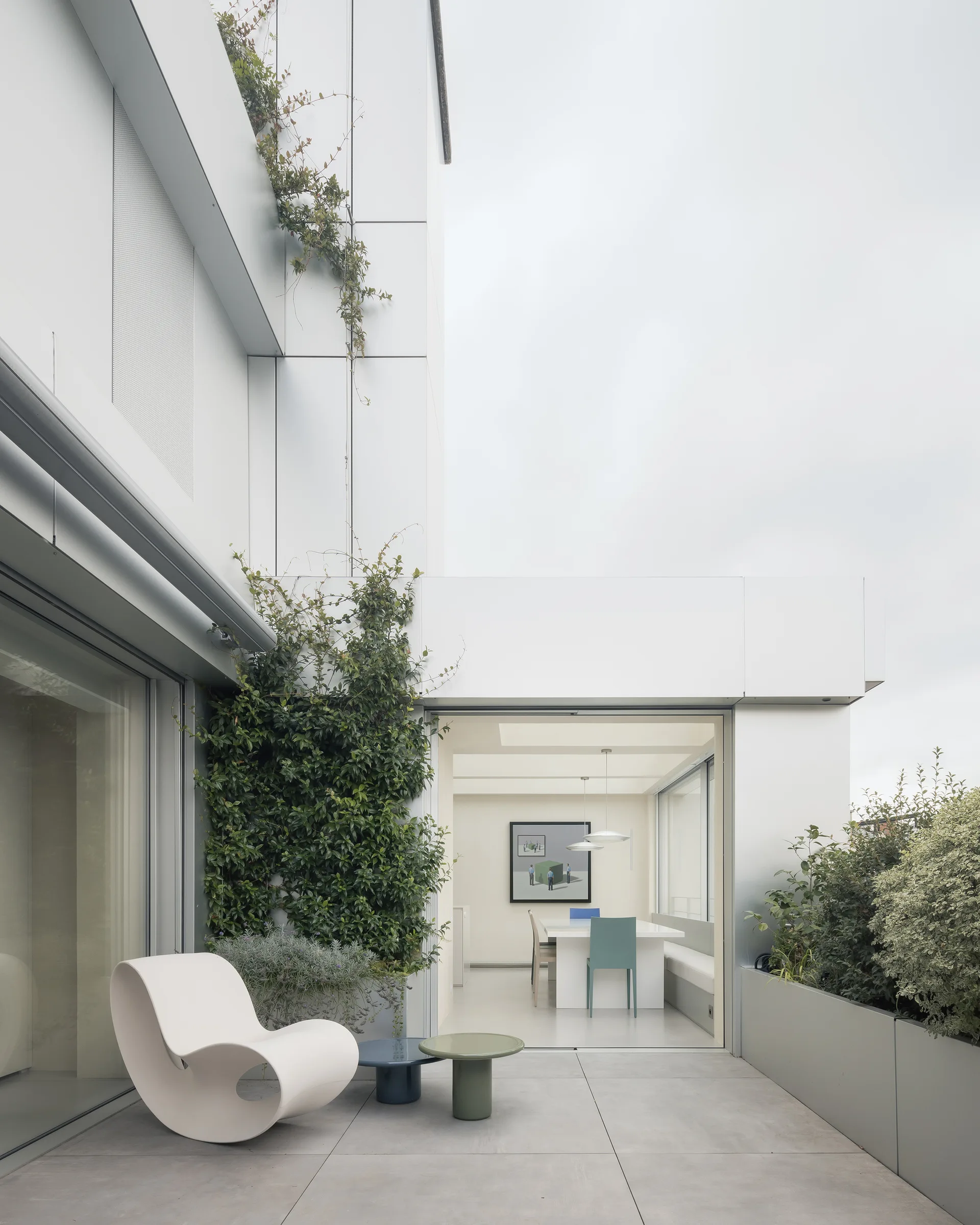 A modern tiled terrace features a white loop-shaped armchair, two colorful side tables, and sliding glass doors opening to an indoor dining space.