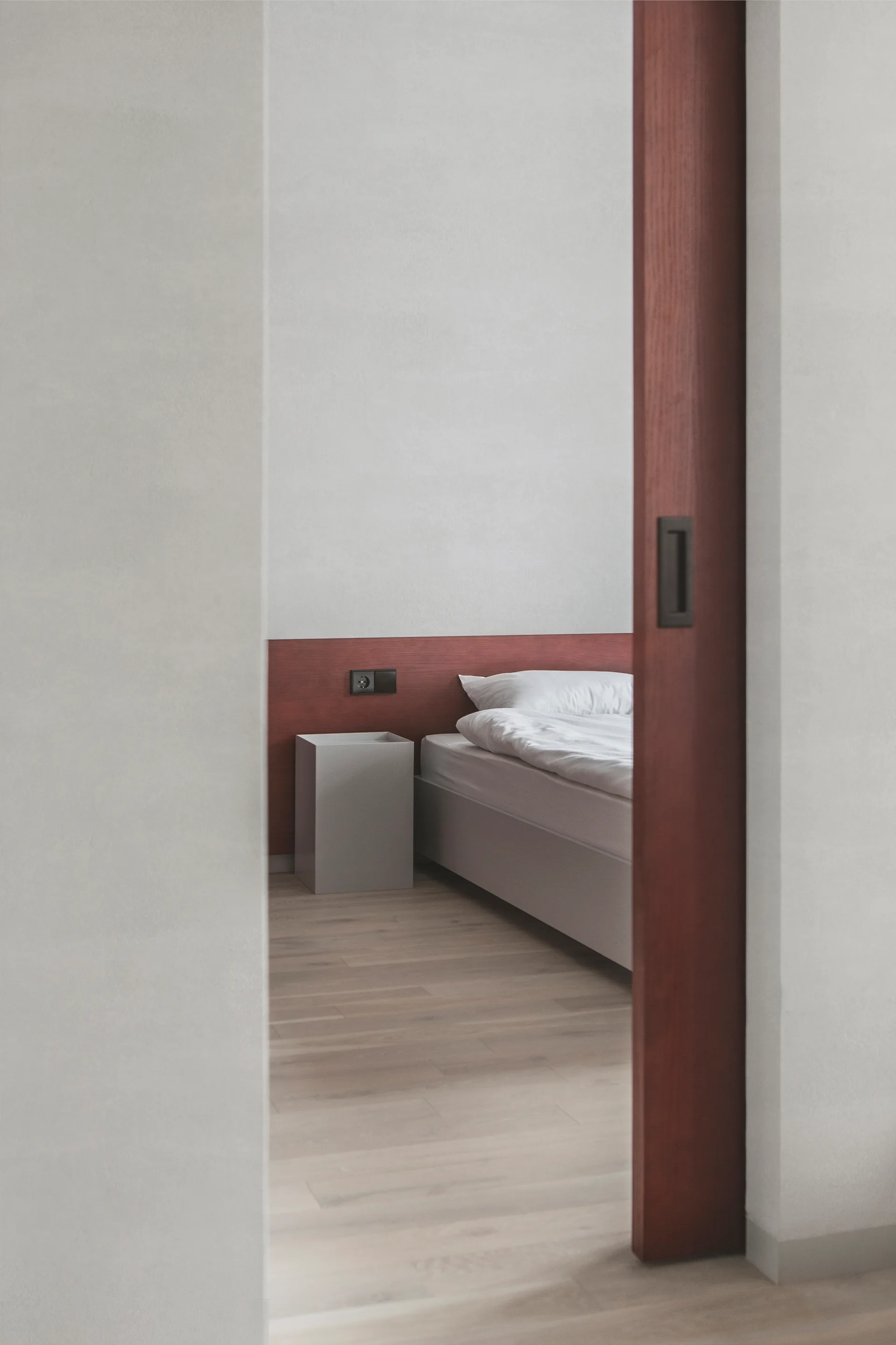 A view through a dark wood sliding doorway into a minimalist bedroom with a low bed, gray cube nightstand, and horizontal wood panel.