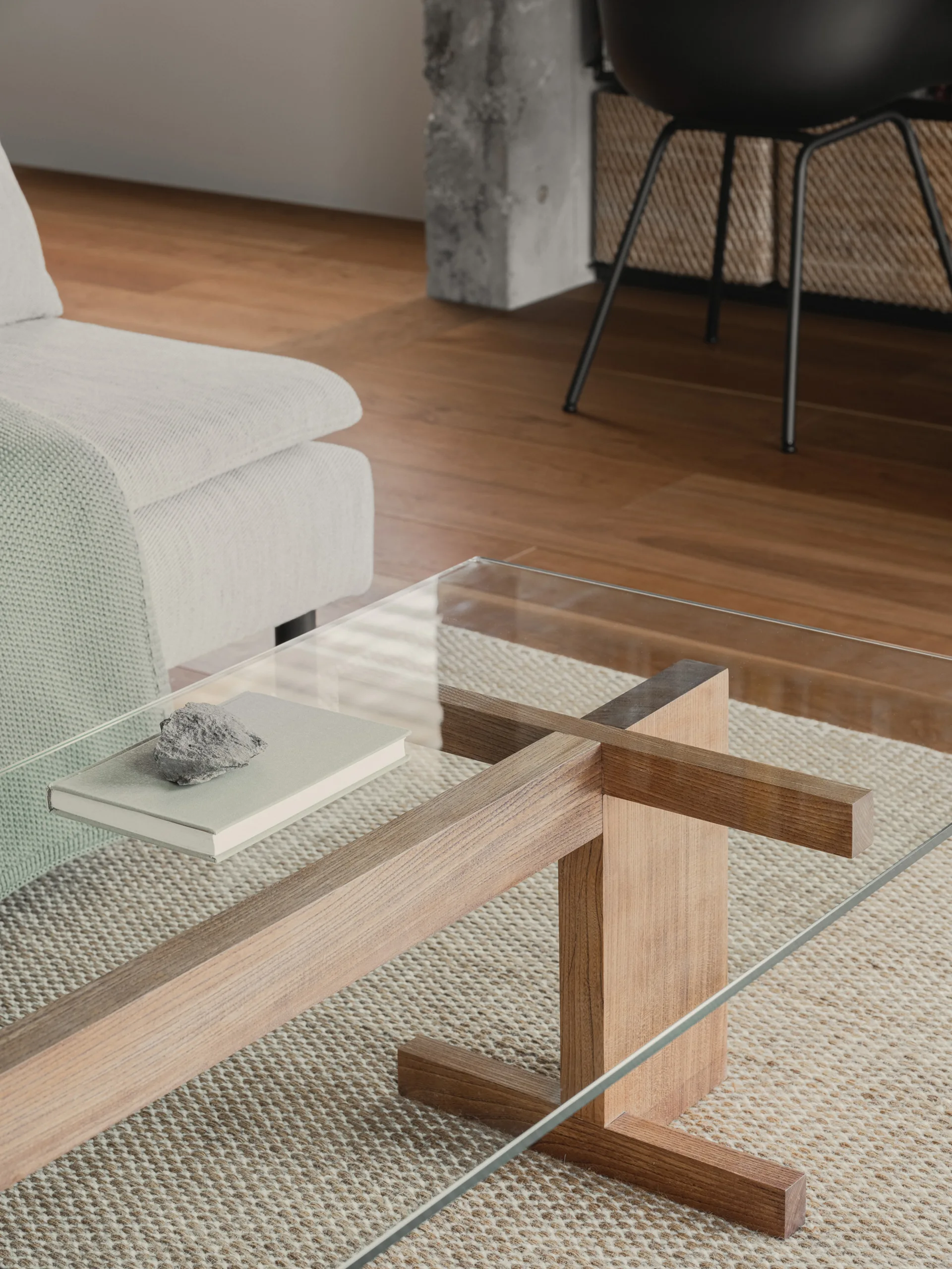 A glass-topped coffee table with a wooden base sits on a textured woven rug near a light grey sofa in a minimalist room.