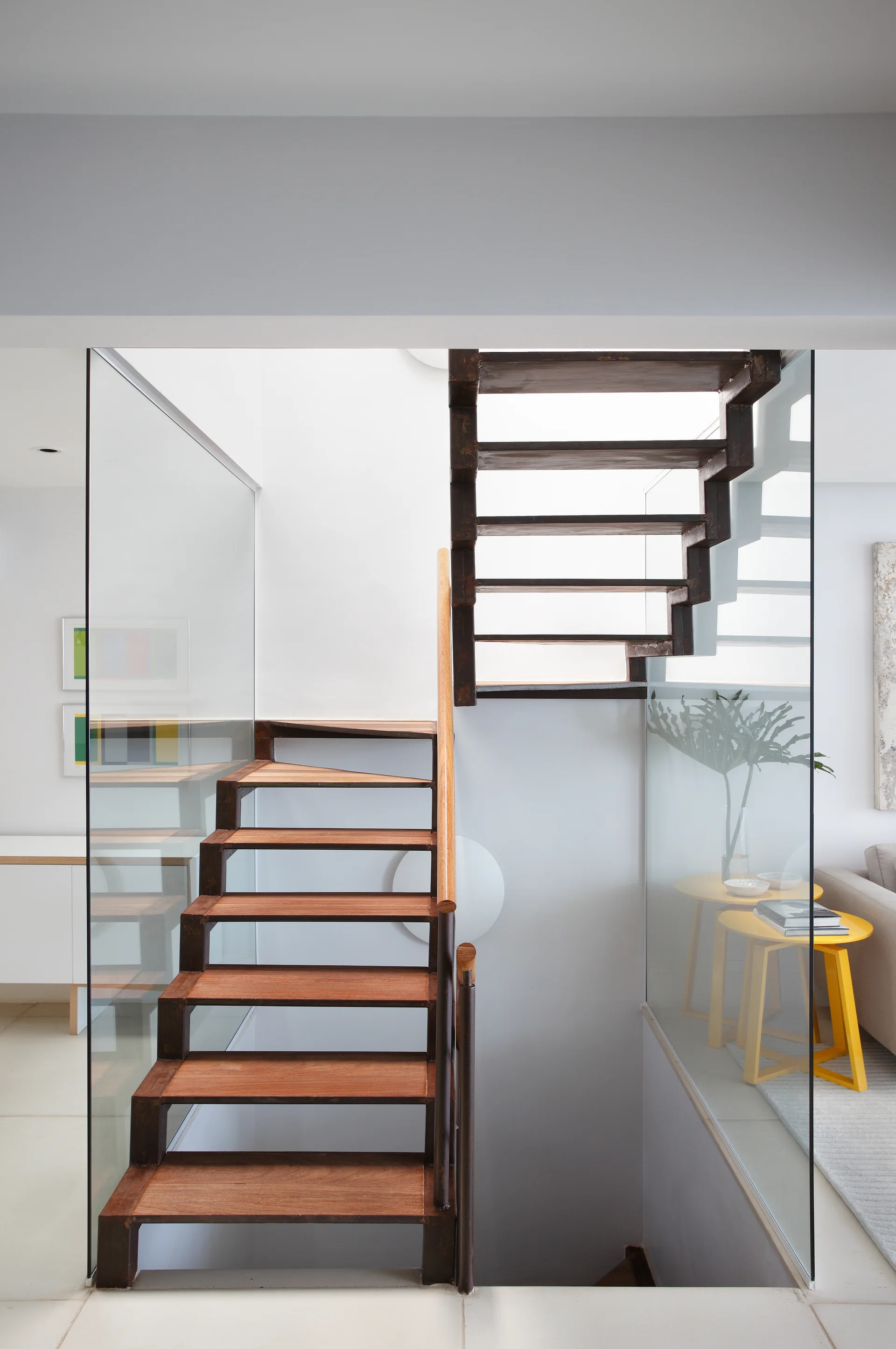 An open-riser wooden staircase with a dark metal frame and a wood handrail is enclosed by floor-to-ceiling glass panels next to a bright living area.