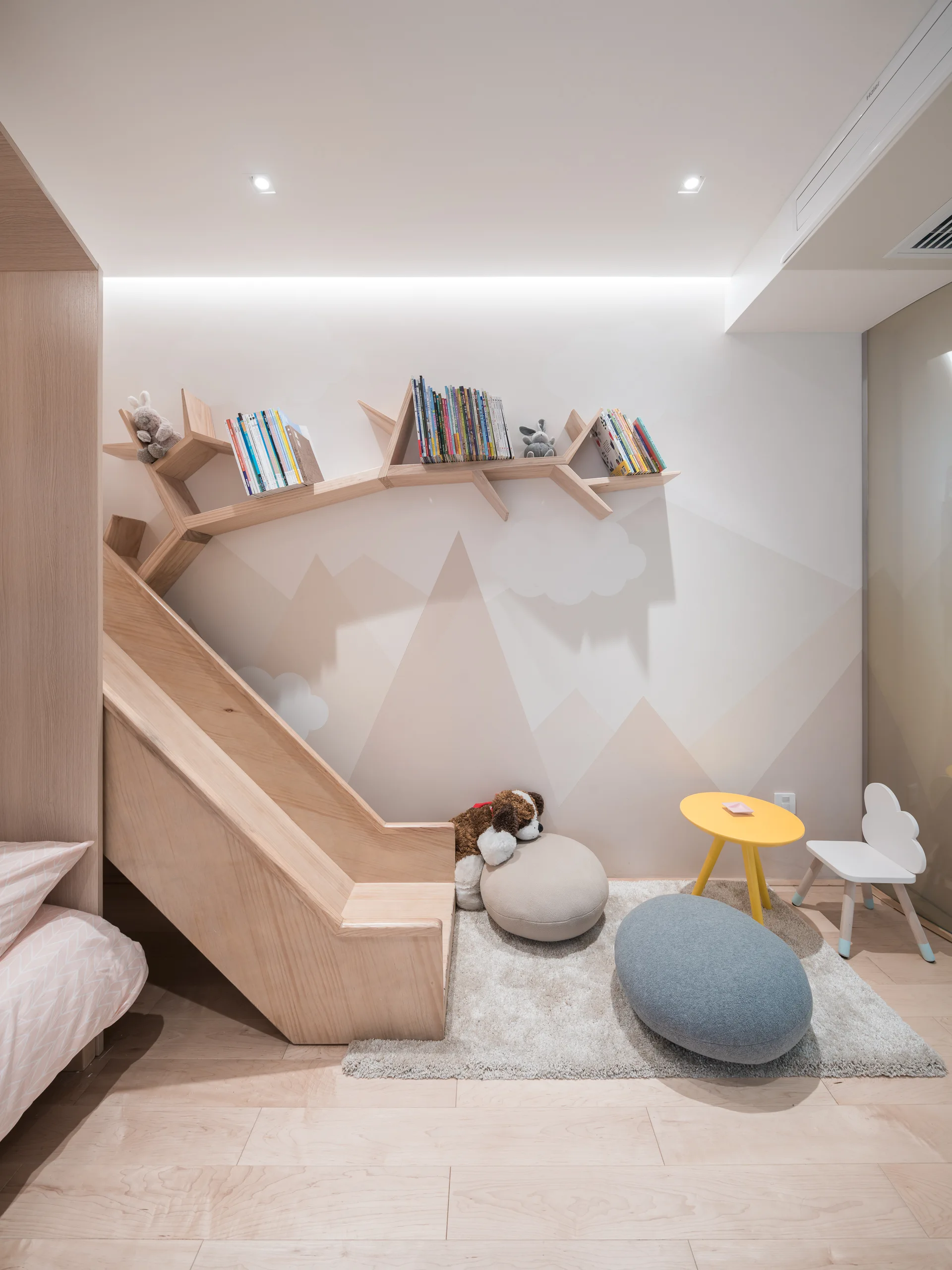 A minimalist kids bedroom featuring a light wood slide, a branch-shaped wall shelf with books, and a mountain mural.