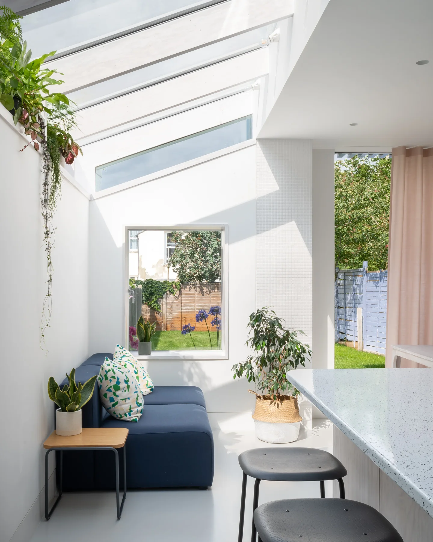 A bright sunroom with a glass roof features a blue sofa, a speckled kitchen island with stools, and green potted plants.
