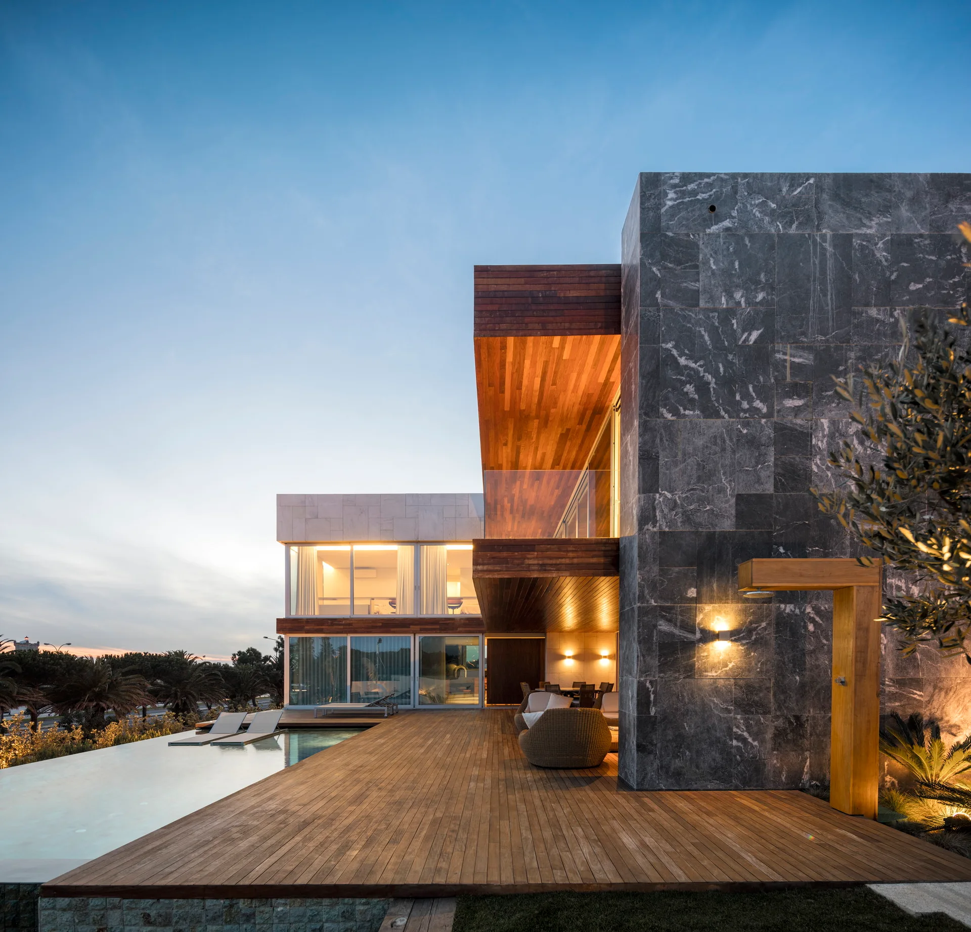 A modern two-story house features a dark grey veined stone facade, wood plank decking, and an infinity pool reflecting the evening sky.