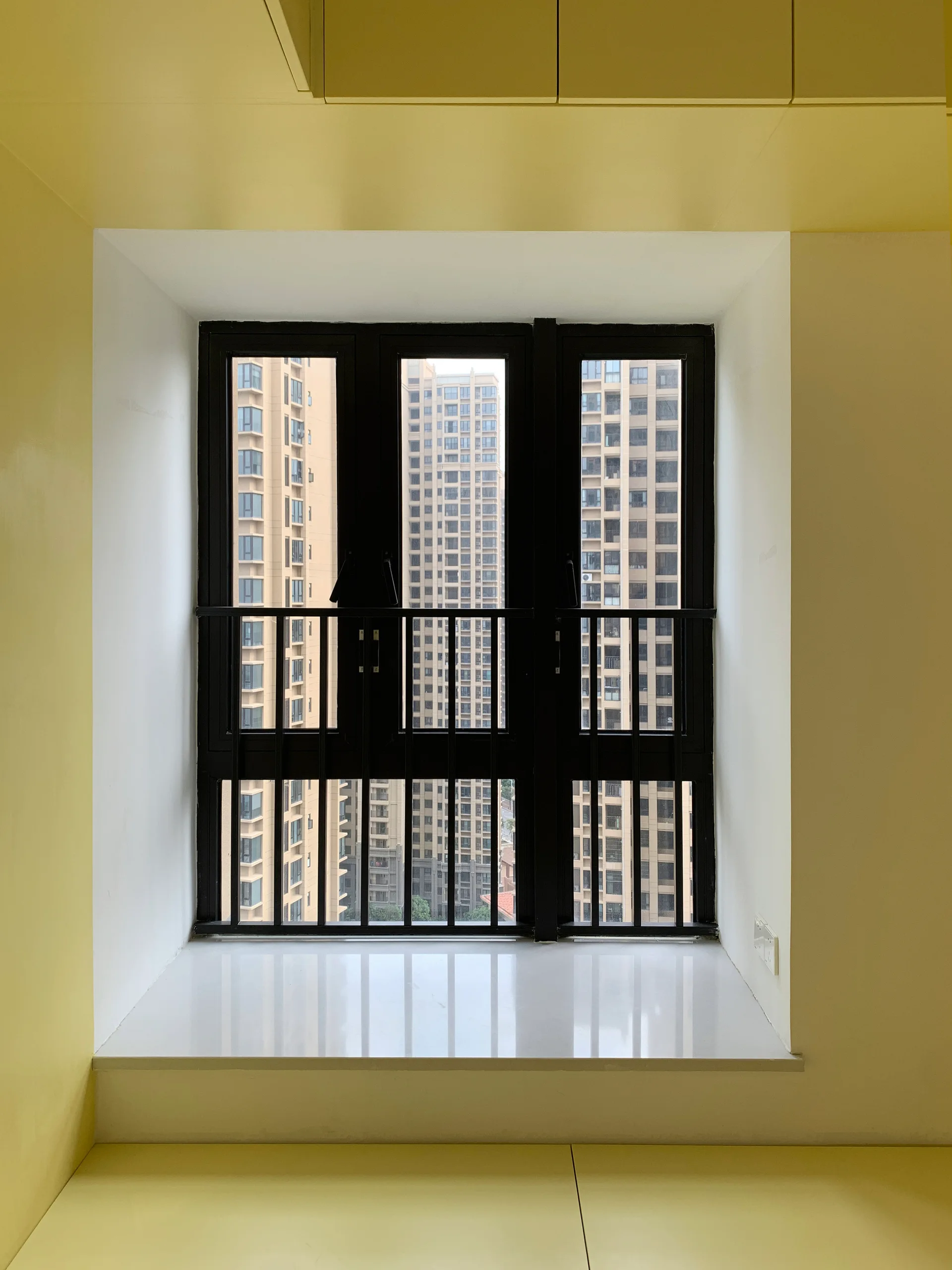A deep-set window with a black metal frame and white glossy sill is surrounded by yellow walls, looking out onto high-rise buildings.