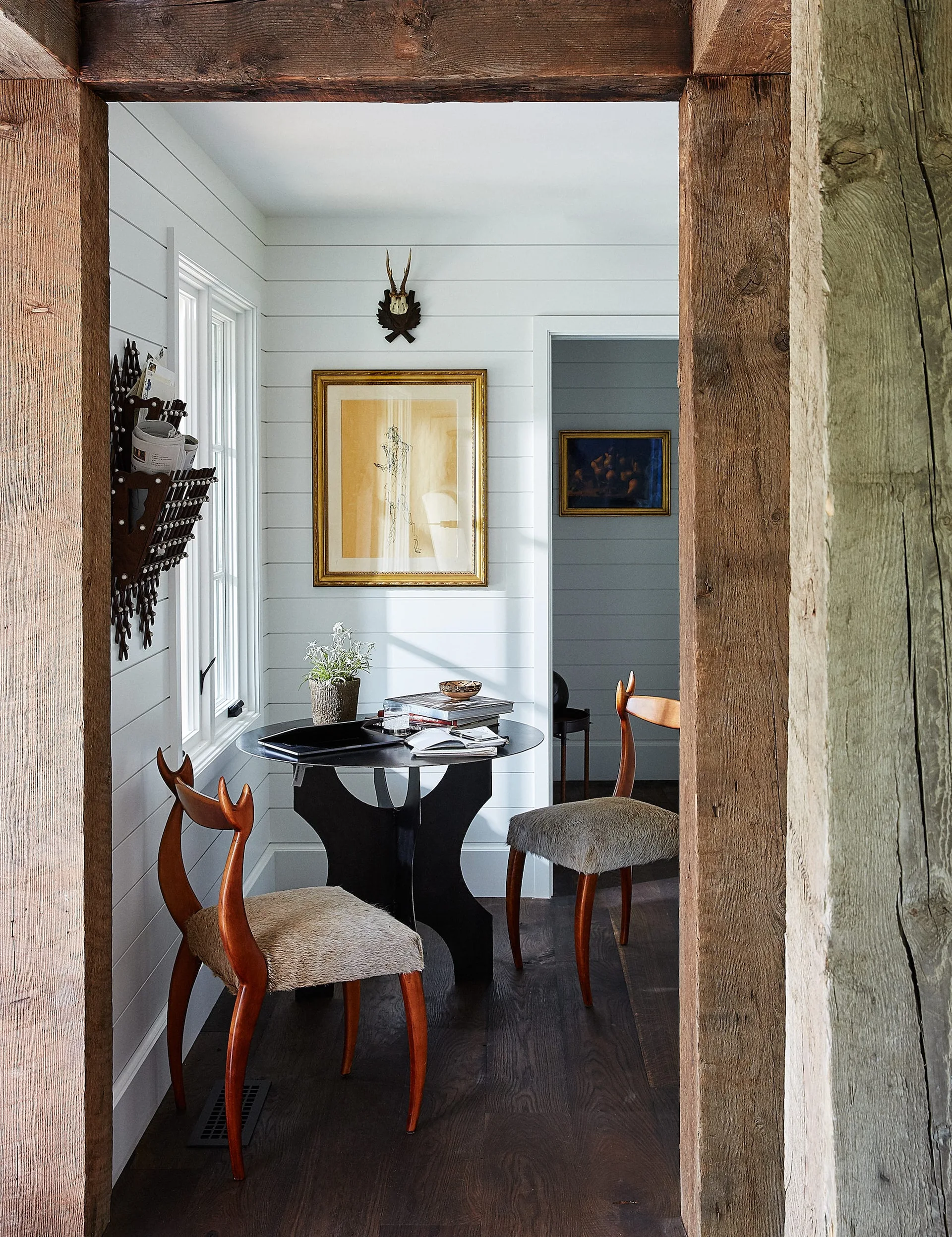 A rustic dining nook viewed through a thick wood doorway features a black table, two chairs with horn-shaped backs, and white shiplap walls.