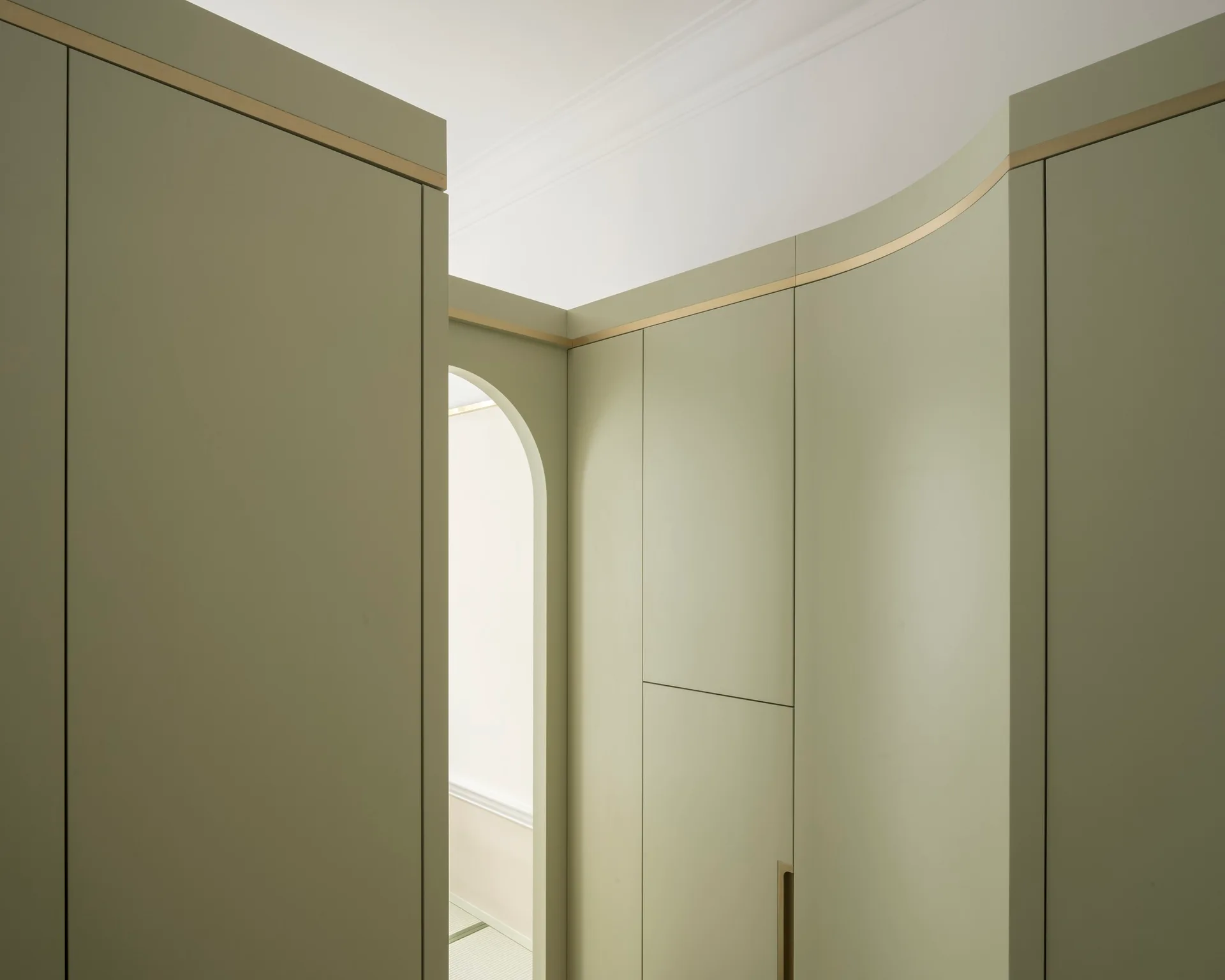 Sleek sage green custom cabinetry curves along a minimalist hallway featuring a gold-toned inlay and an arched opening leading to another room.