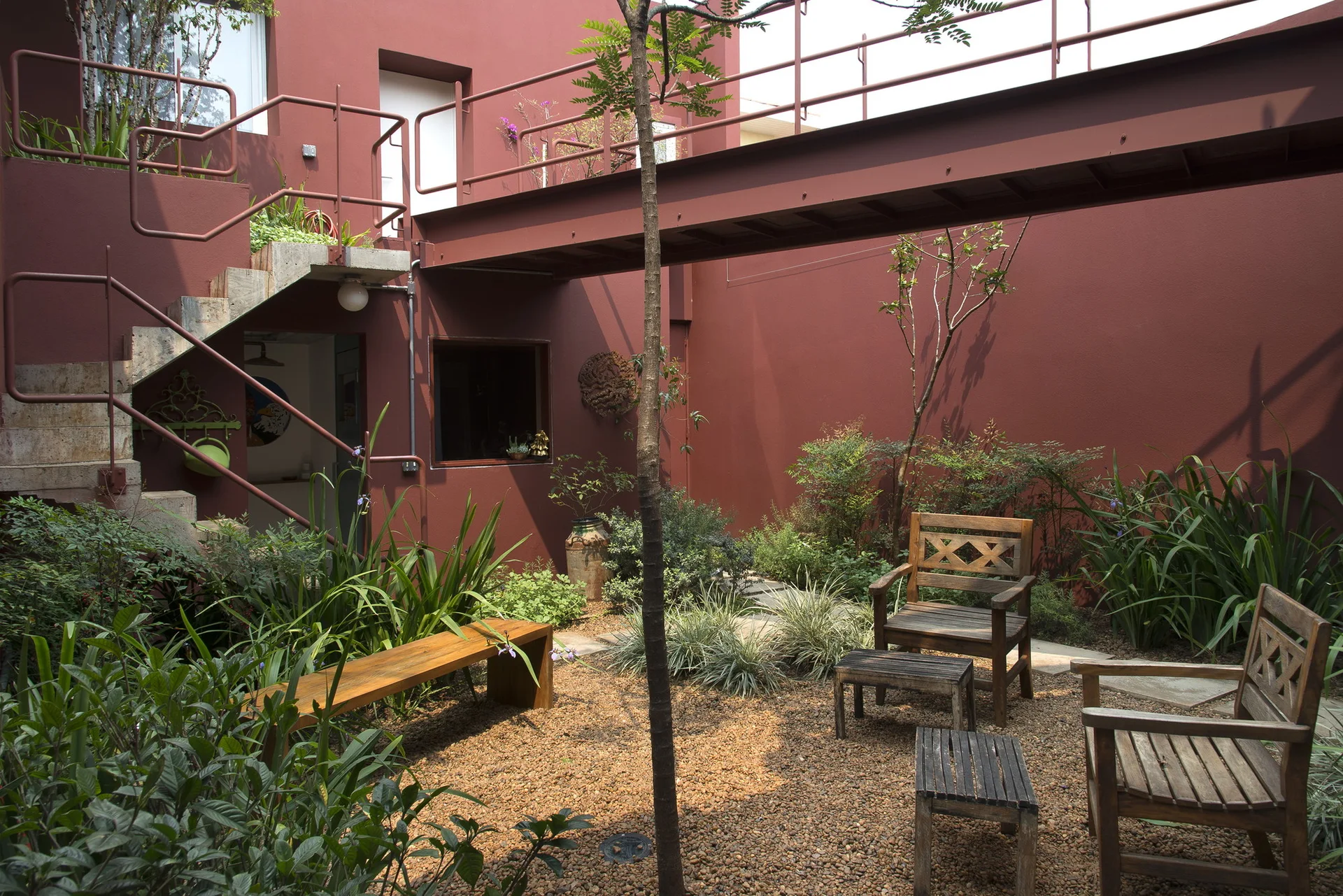 An enclosed outdoor courtyard featuring dark red painted walls, a metal footbridge overhead, and simple wooden chairs on a gravel ground with lush green plants.