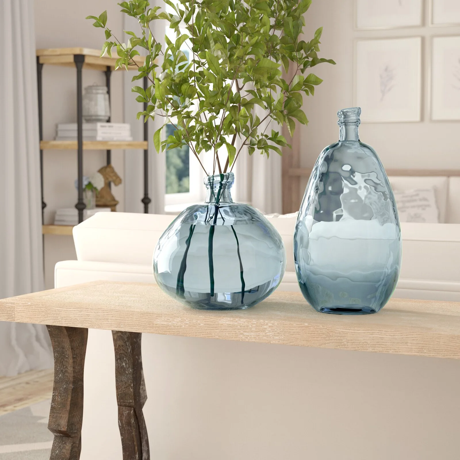Two blue tinted glass vessels sit on a light wood console table decorated with green branches in a bright room.