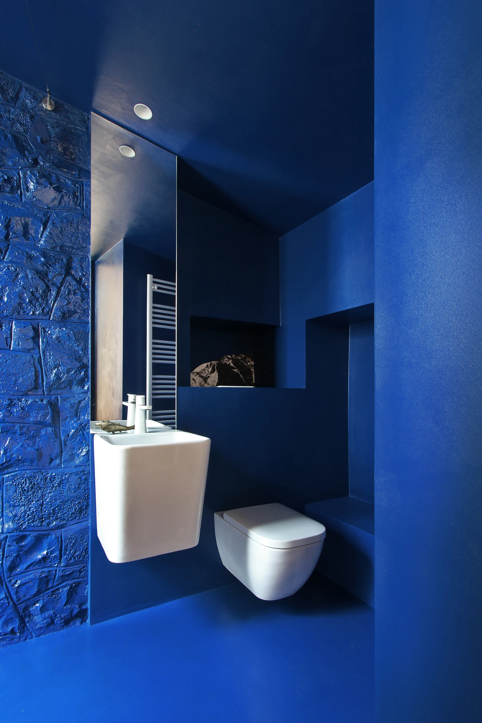 A minimalist powder room with monochromatic blue walls, ceiling, and textured stone, featuring a white wall-mounted toilet and square floating sink.