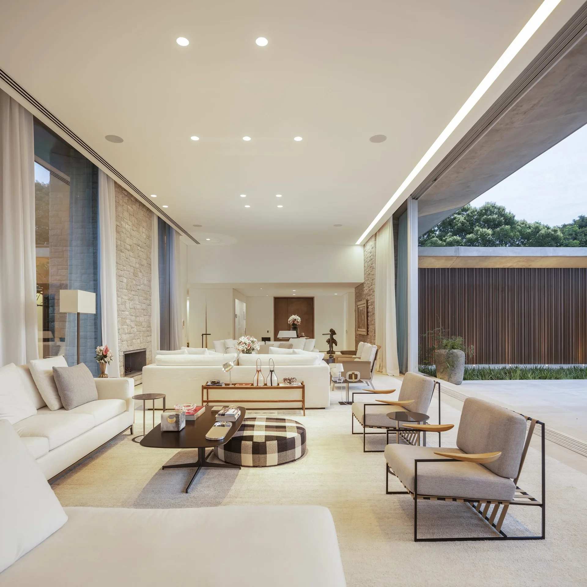 A spacious white living room features low sofas, a checkered ottoman, stone accent walls, and a large sliding glass wall opening to a terrace.