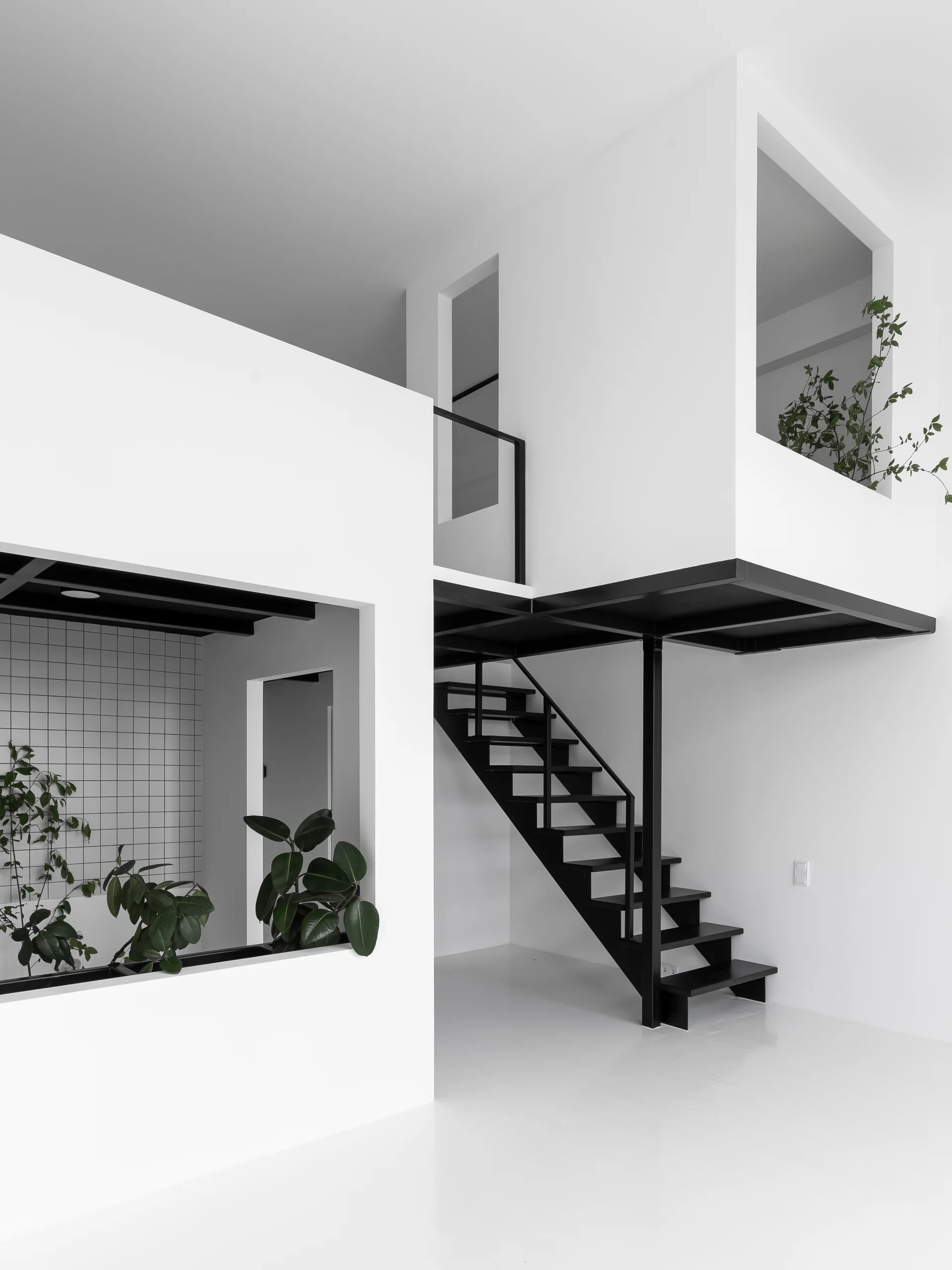 A minimalist white loft interior features a black metal staircase leading to a mezzanine, alongside geometric wall openings and potted green plants.