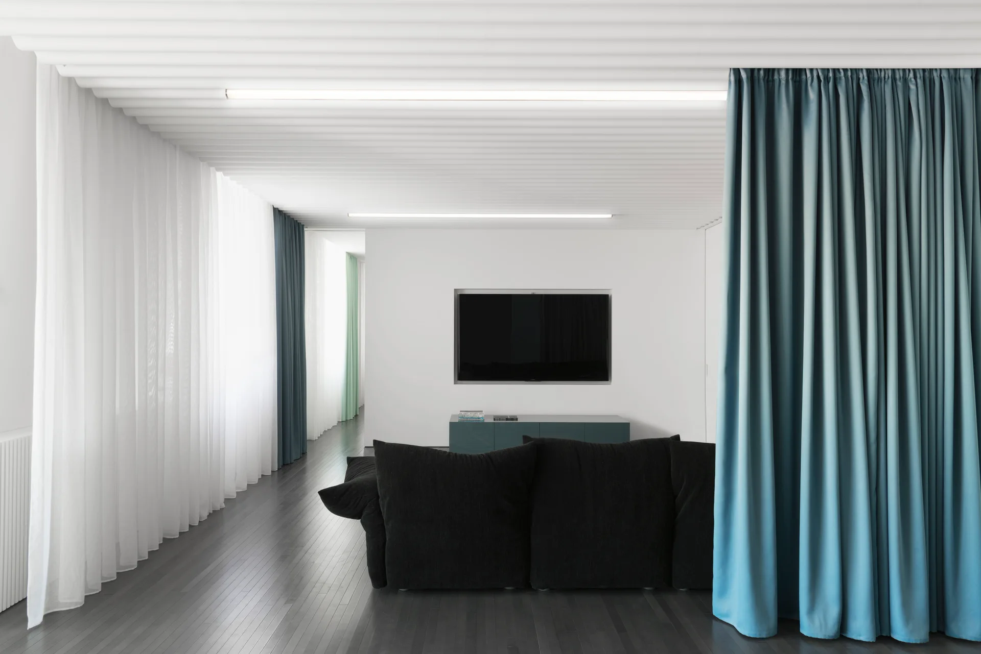 A minimalist living room features a dark sofa, a slatted white ceiling, and long blue and sheer white curtains over dark wood floors.