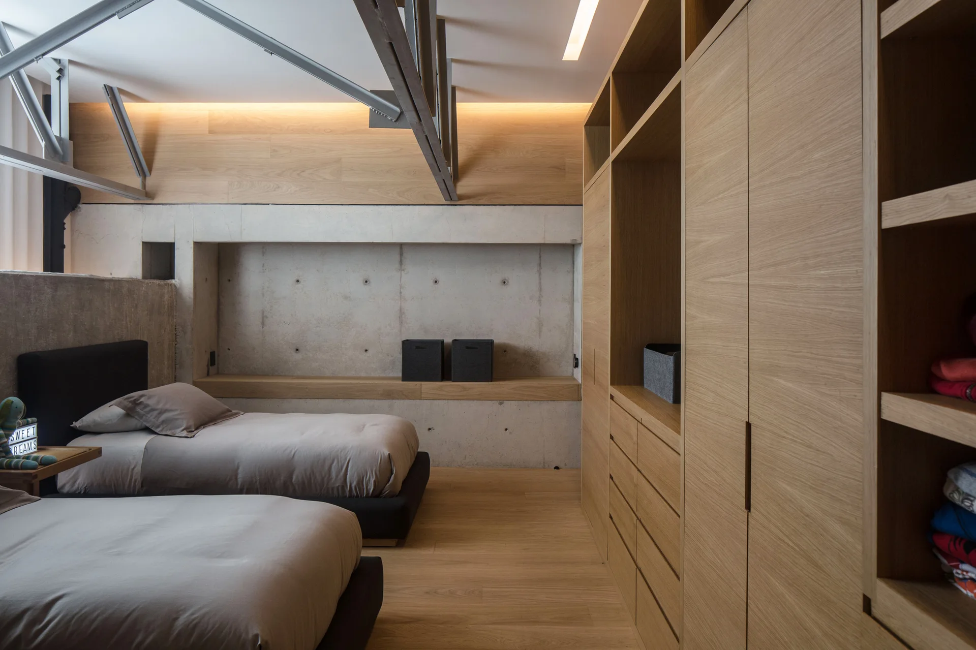 A modern minimalist bedroom featuring twin low platform beds, an exposed concrete wall with a recessed niche, and a large built-in oak wardrobe.