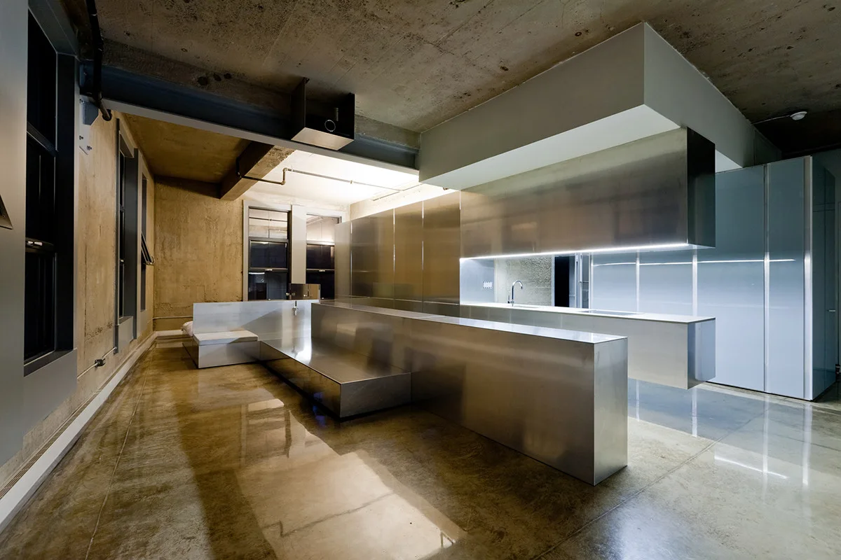 A minimalist industrial kitchen featuring extensive brushed stainless steel cabinetry, a large metallic island, and polished concrete floors and ceiling.