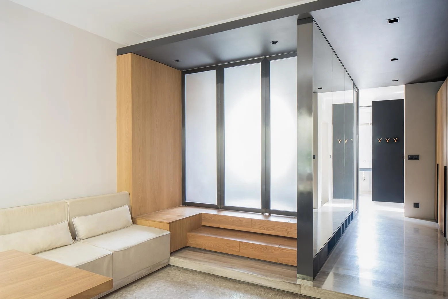 A minimalist living area features a light beige sofa next to a raised wooden platform and steps beneath frosted glass windows and a glass partition hallway.