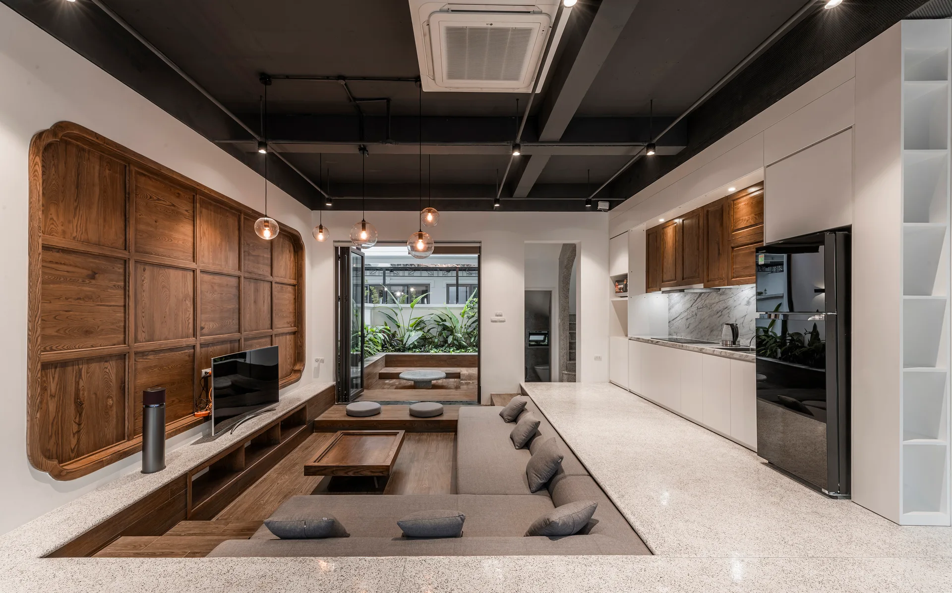 A modern open-concept space features a sunken grey sectional sofa, a wood-paneled TV wall, and an integrated white kitchen with terrazzo flooring.