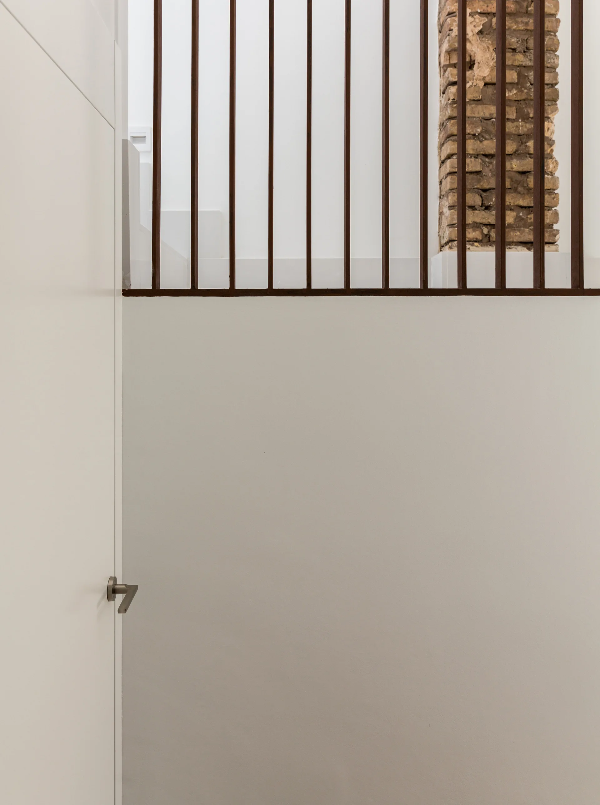 A minimalist dark metal staircase railing stands above a smooth off-white wall next to a vertical exposed brick structural column.