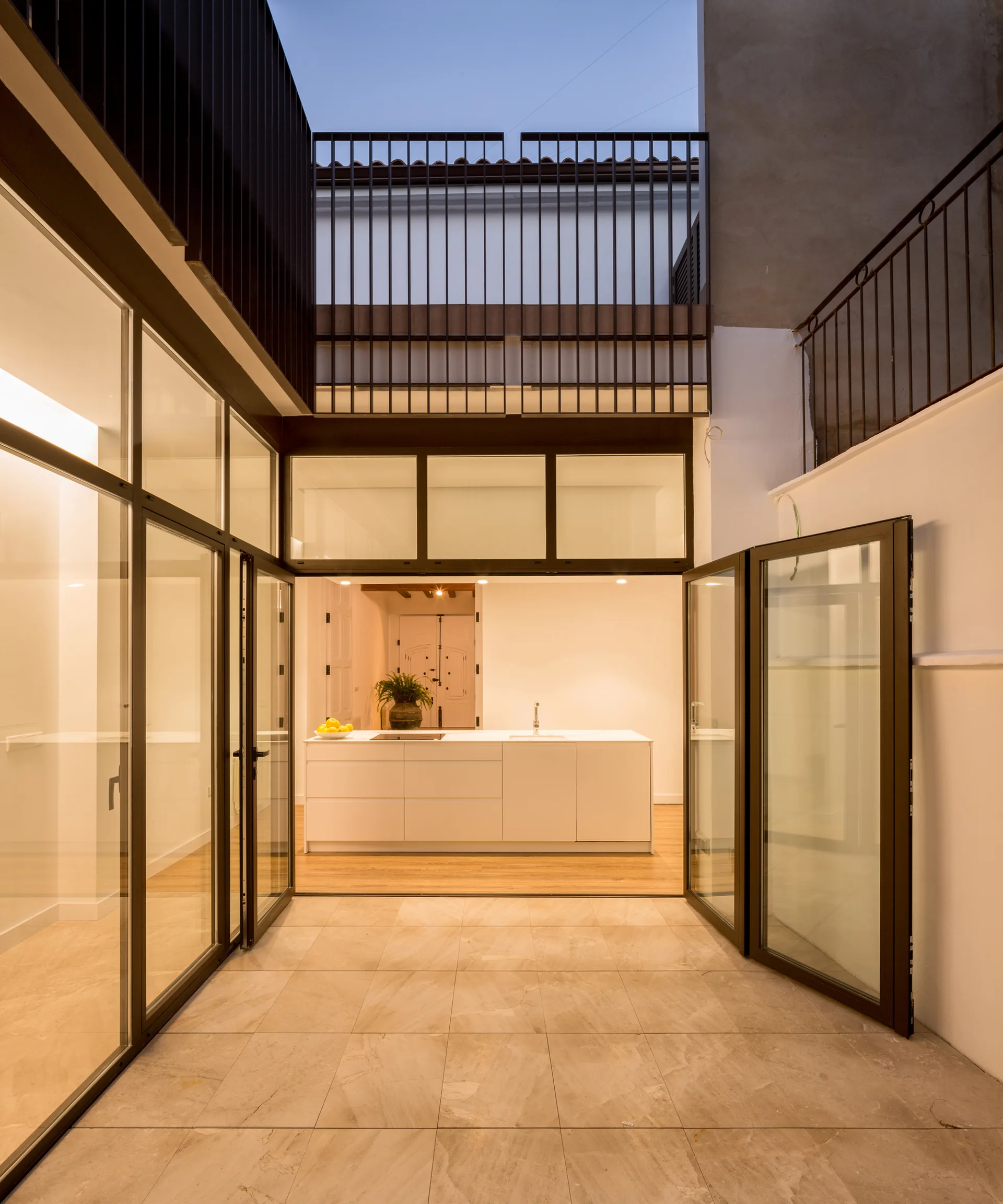 Large glass doors open from a tiled outdoor courtyard into a brightly lit minimalist white kitchen with a central island.