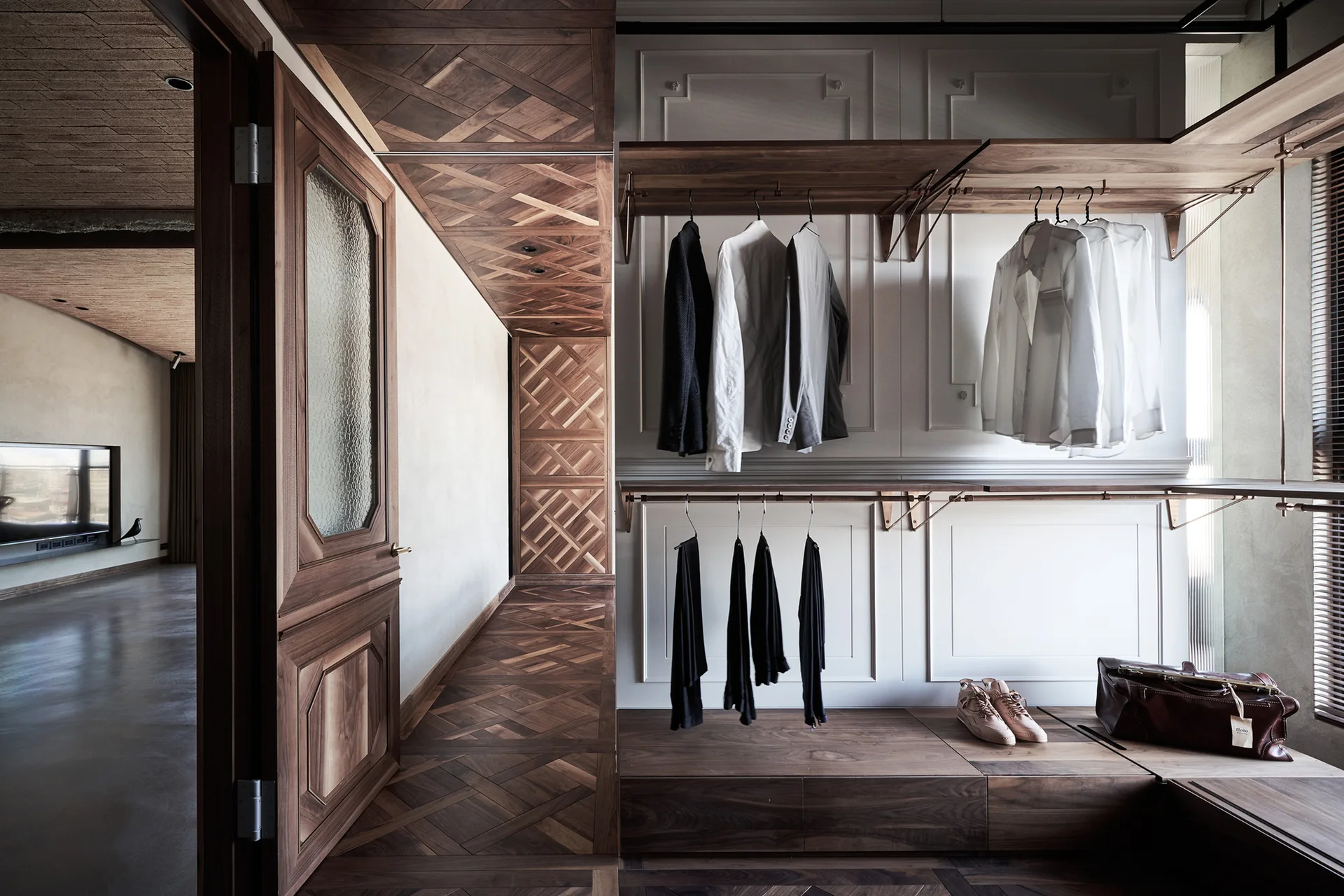 An open wardrobe with dark wood shelves and hanging clothes is set against white paneled walls next to a patterned wood parquet floor.