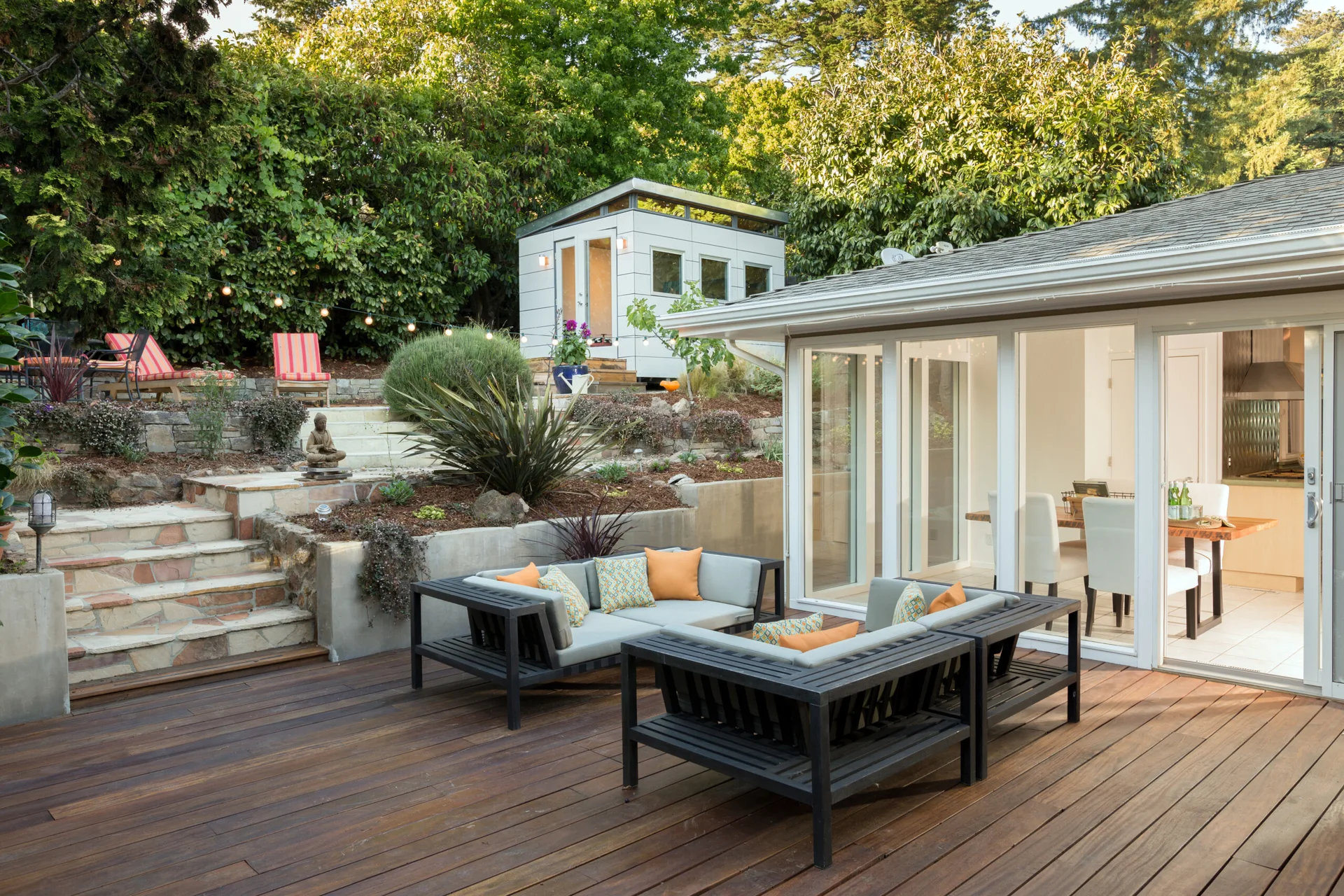 An outdoor wood deck features a grey sectional sofa with orange pillows, set against a tiered garden with stone steps and a white studio shed.