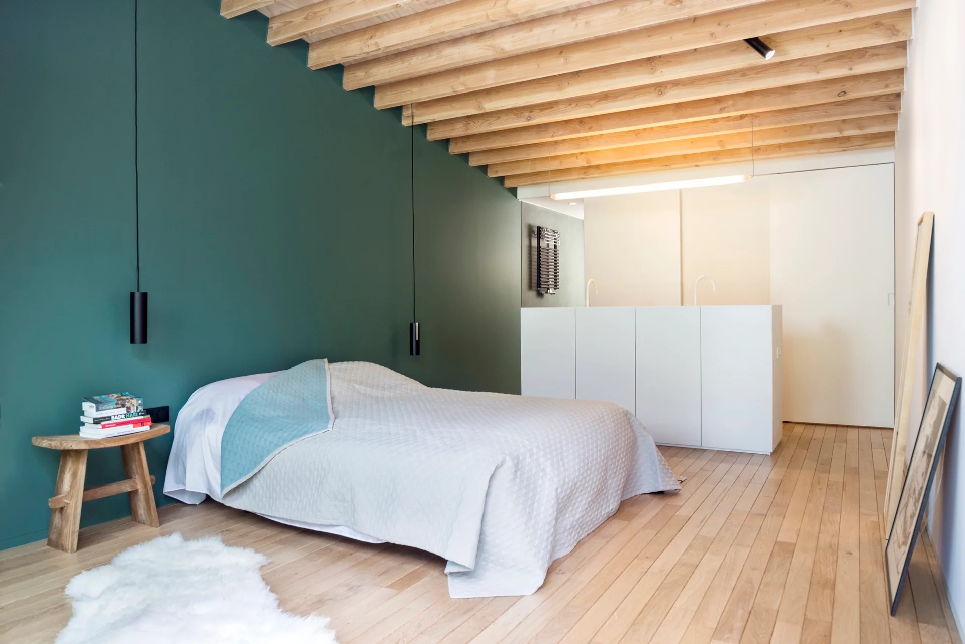 A minimalist bedroom features a dark green accent wall, low bed, exposed wood ceiling beams, and light-colored hardwood flooring.