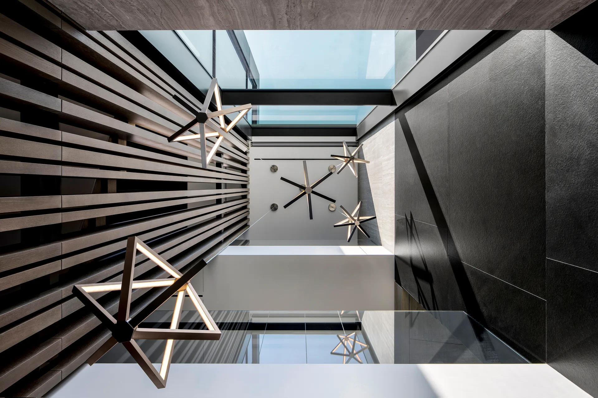 An upward perspective of a modern atrium showing geometric star-shaped pendant lights, a vertical wood slat wall, and a large overhead skylight.