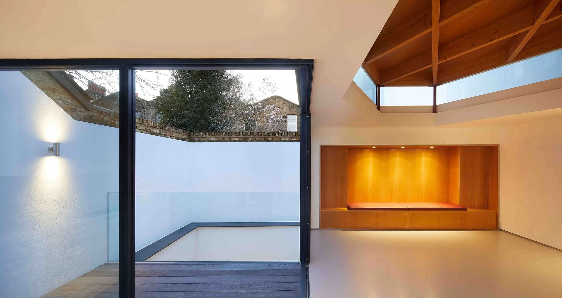 A minimalist living space features a vaulted timber ceiling, a wooden built-in bench with a red cushion, and sliding glass doors opening to a sunken patio.