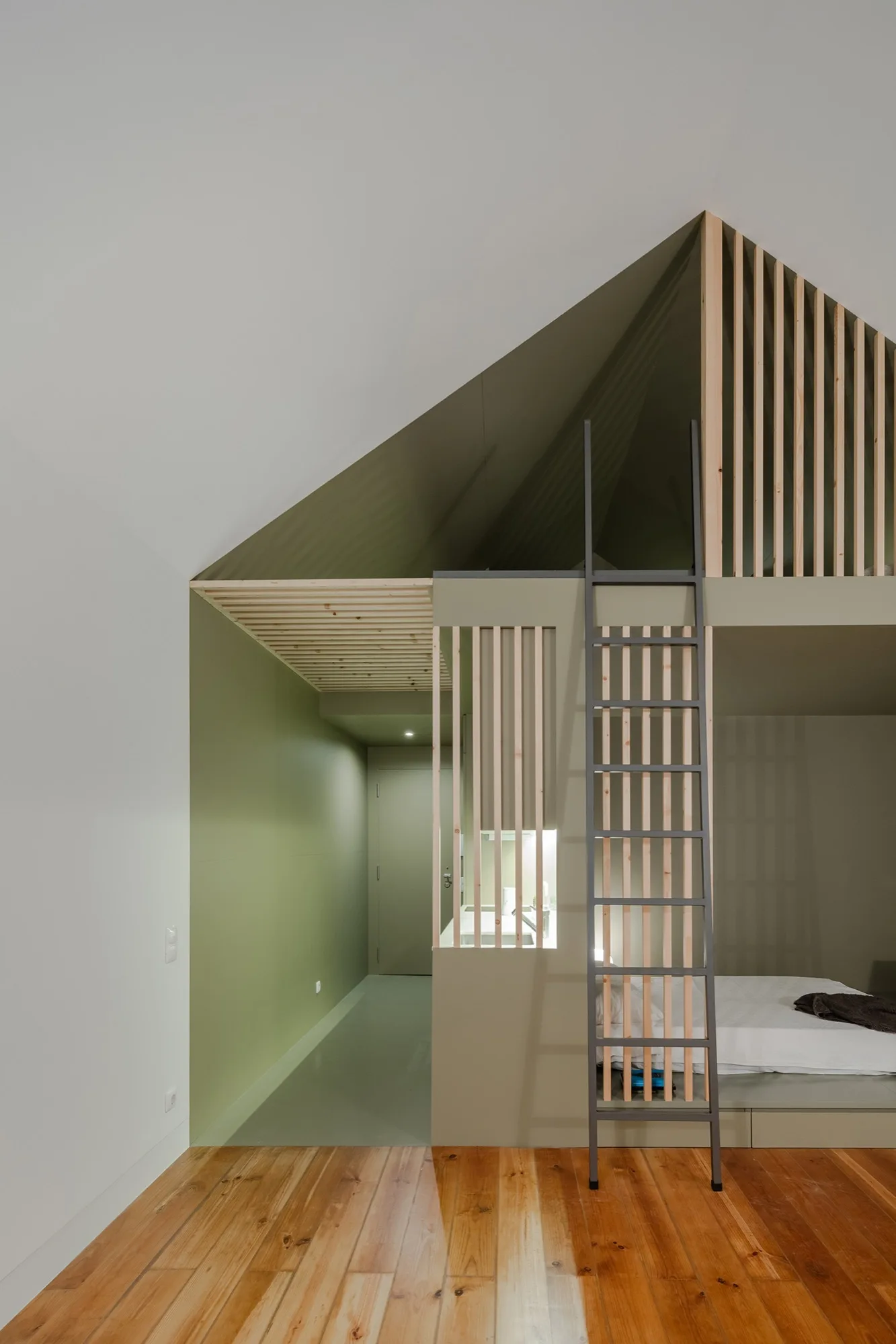 A minimalist bedroom space featuring a sage green built-in loft bed structure with a vertical wooden slat screen, a dark ladder, and wood flooring.