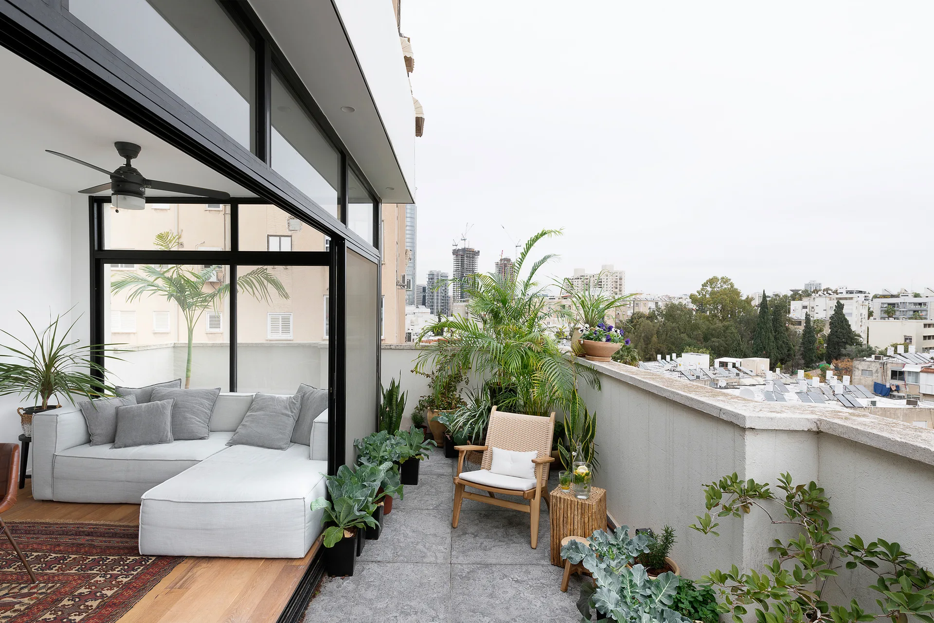 An urban rooftop terrace features a grey sectional sofa inside a glass enclosure and a woven wood armchair surrounded by potted plants.