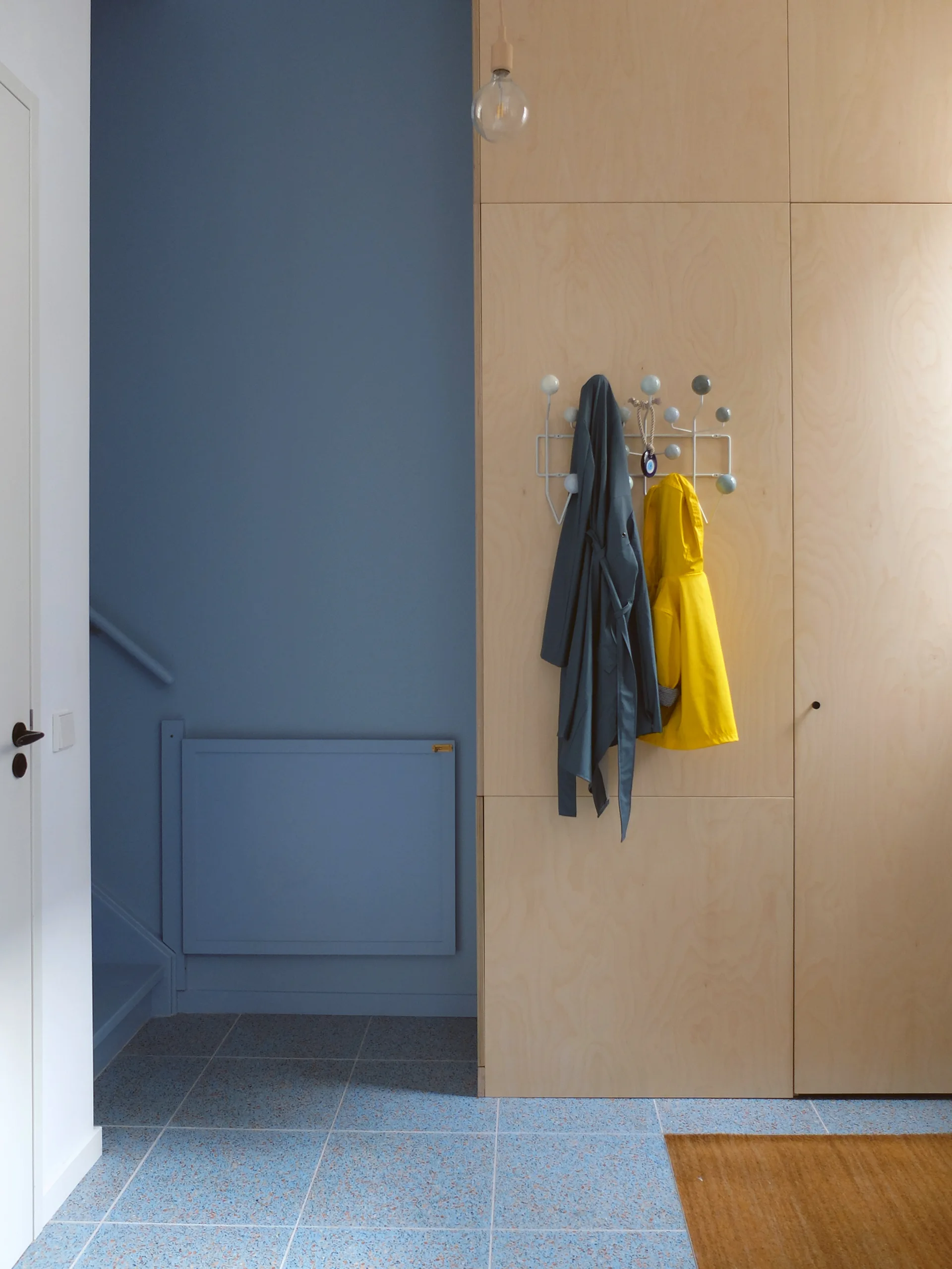 A minimalist entryway features full-height plywood cabinetry with a white coat rack, a solid blue painted wall, and blue speckled terrazzo tile flooring.