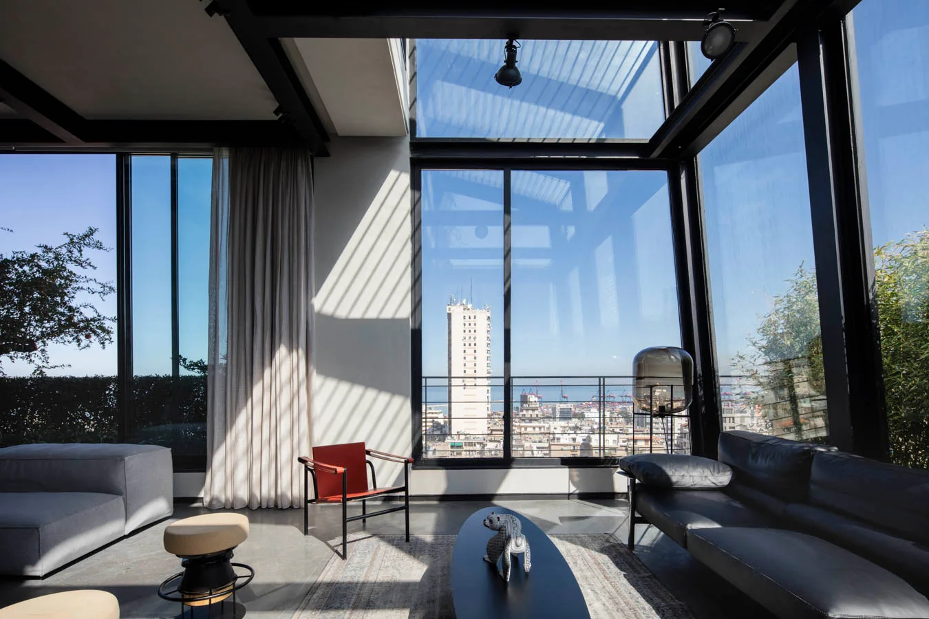 A modern living room features floor-to-ceiling windows showing a city view, black ceiling beams, a dark leather sofa, and a red accent chair.