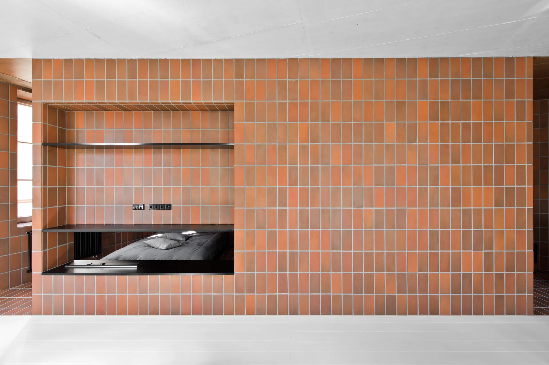 A modern bedroom features a large wall clad in vertical orange terracotta tiles with a built-in recessed sleeping alcove containing a low bed.