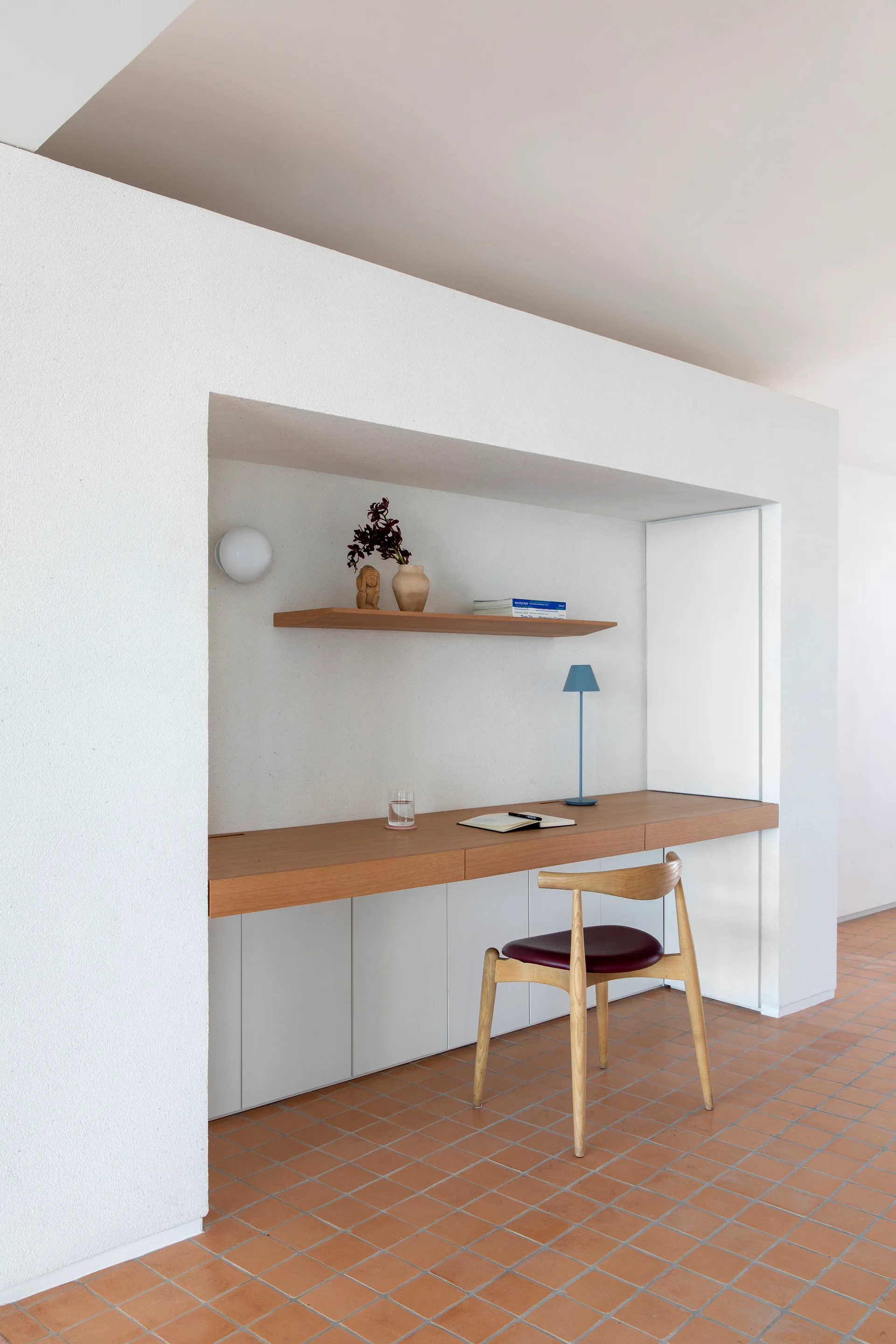 A minimalist wooden desk built into a white plaster wall alcove above warm terracotta floor tiles.