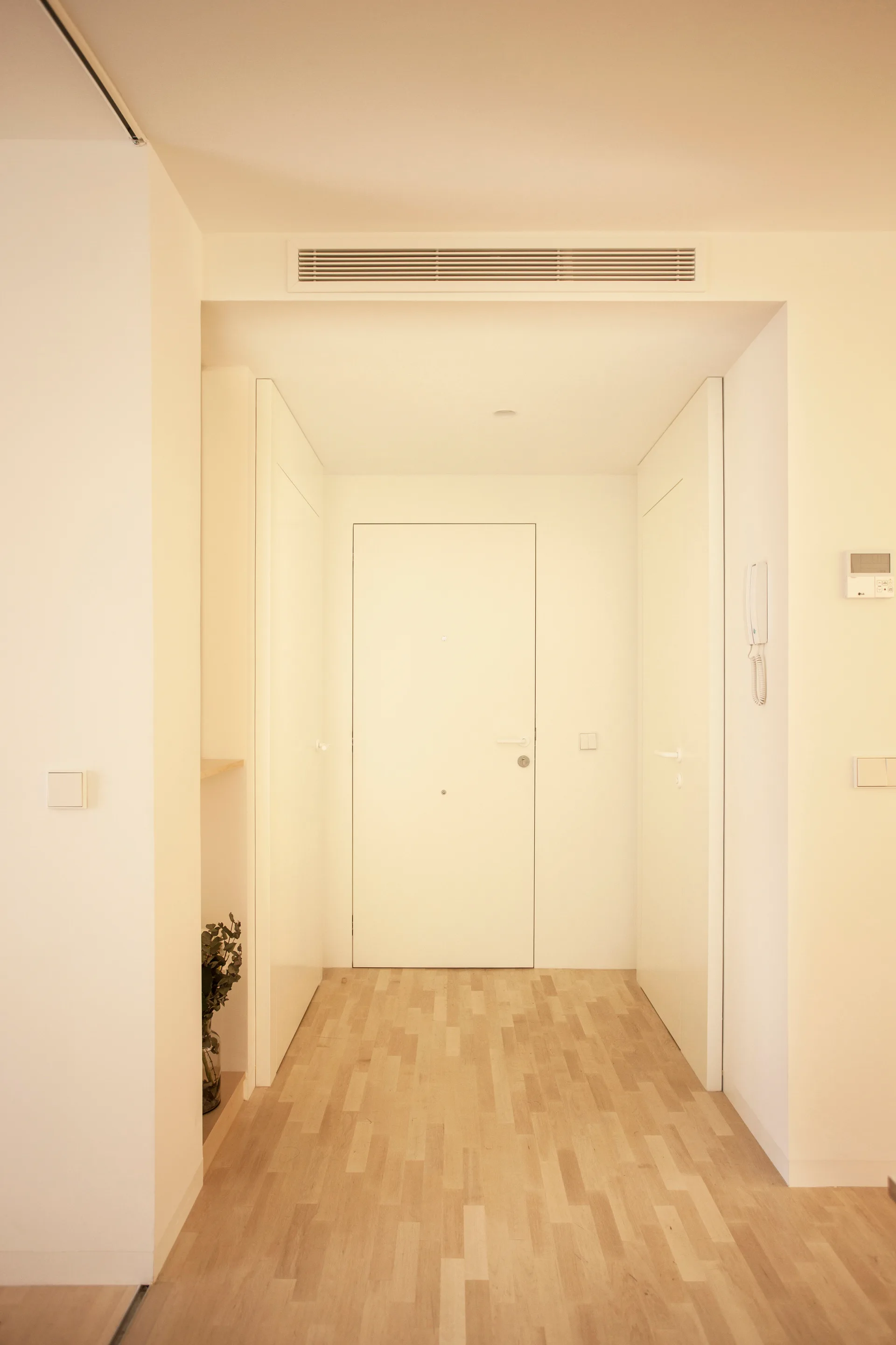 A minimalist white entryway featuring seamless flush doors, light wood plank flooring, and a linear ceiling vent.