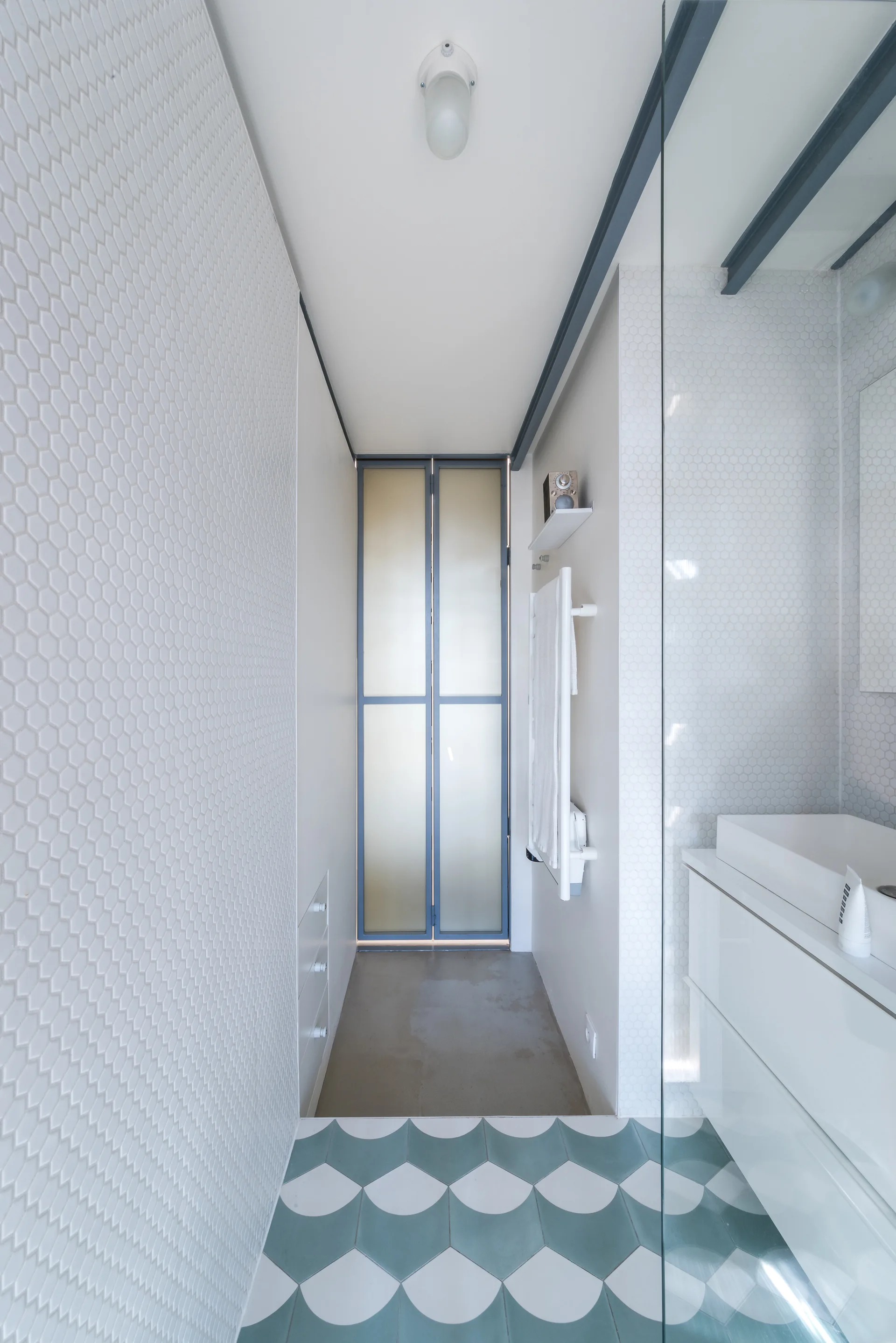 A narrow modern bathroom with a scalloped teal floor, a white hexagonal mosaic tile wall, a minimalist vanity, and a frosted glass bifold door.