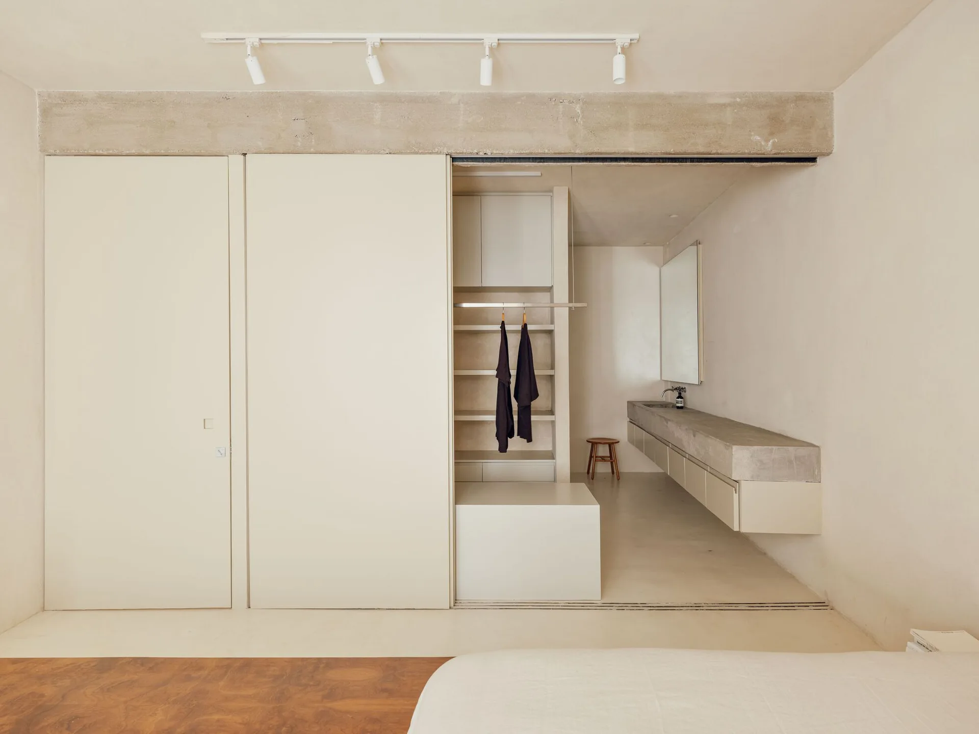A minimalist bedroom dressing area with seamless beige sliding closet doors, an exposed concrete beam, and a long floating stone vanity.