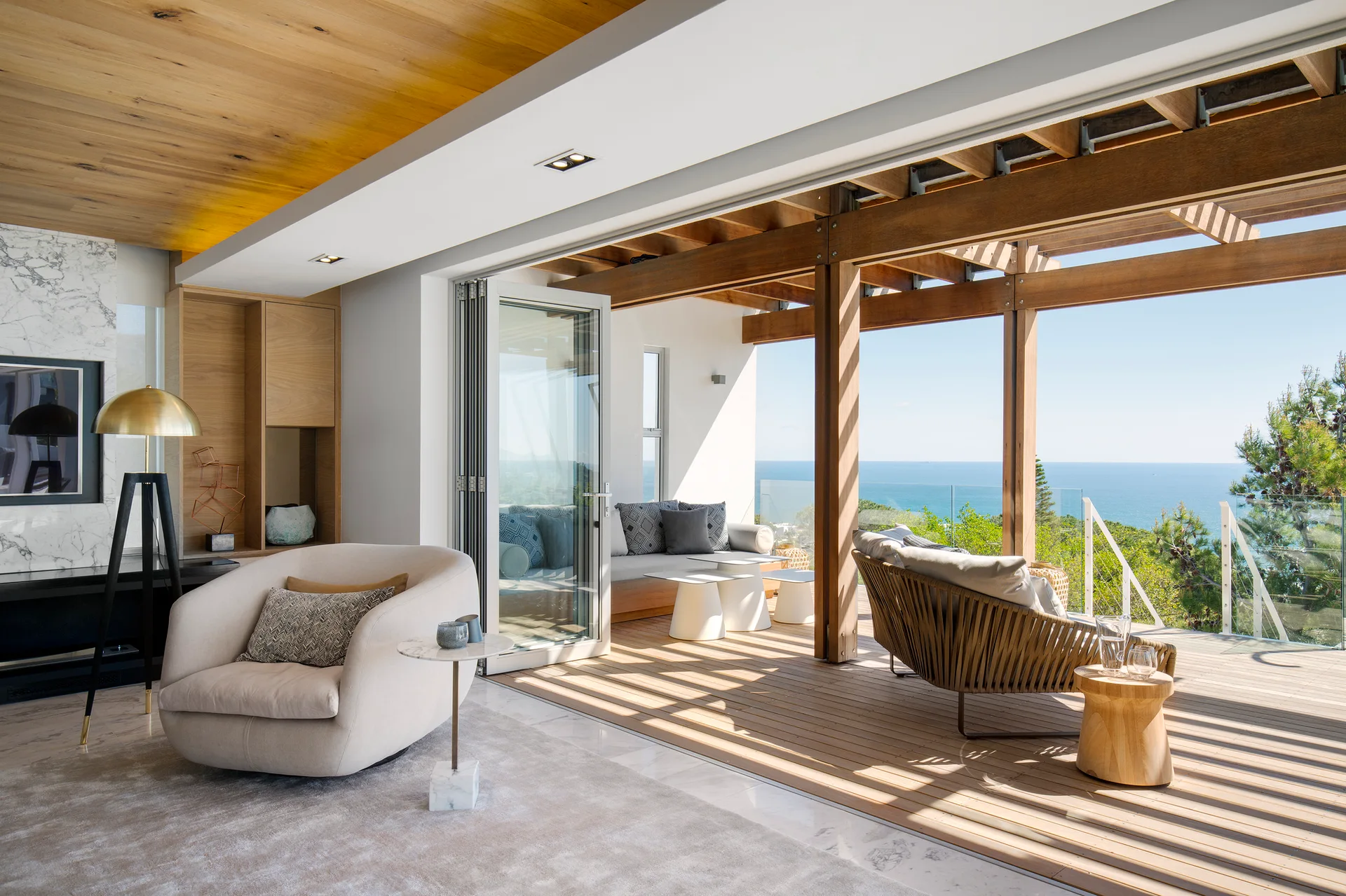 A modern living room with a marble floor and wood ceiling transitions through open bi-fold doors to a wooden terrace overlooking the ocean.