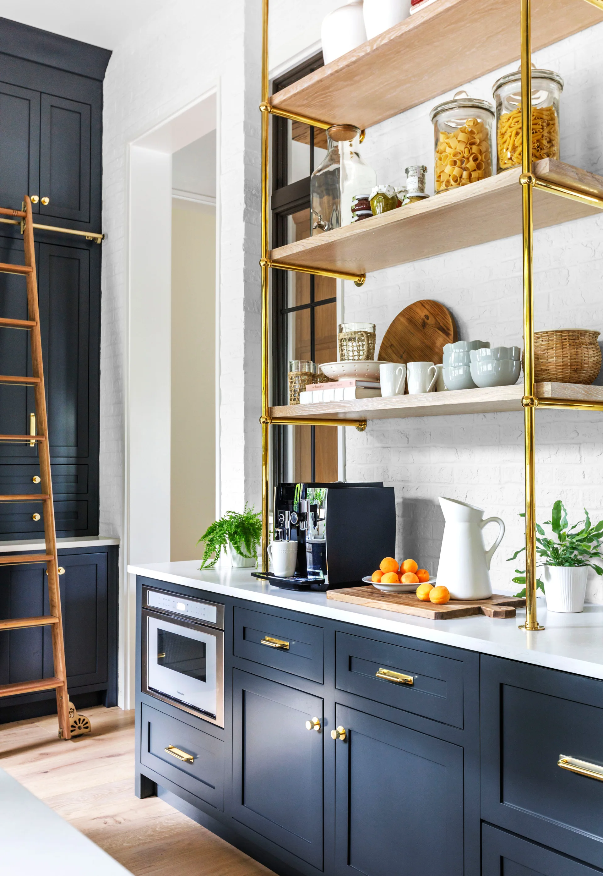 Navy blue kitchen cabinets feature gold hardware, white countertops, and open wood shelving with a brass frame against a painted white brick wall.