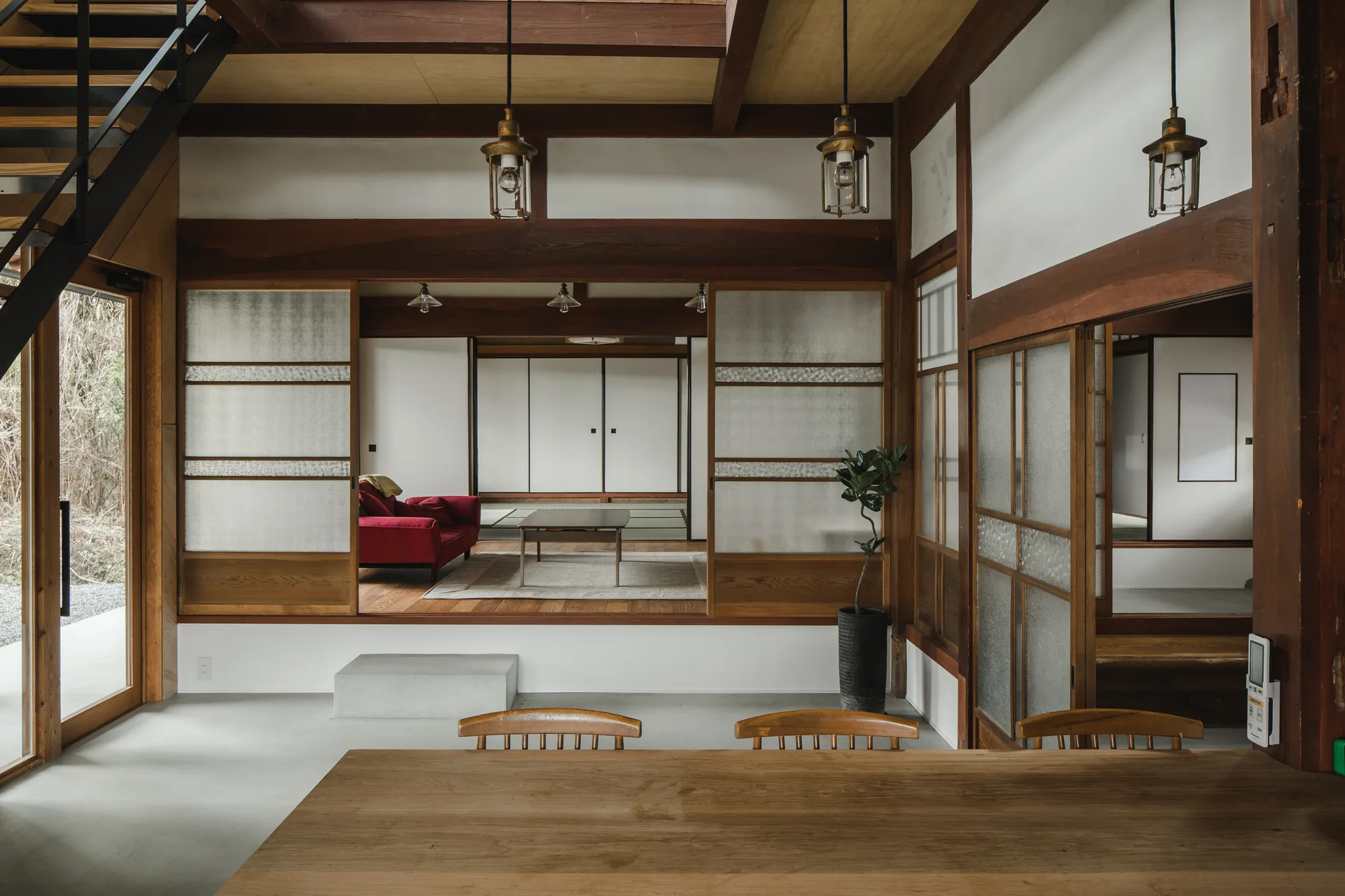 A modern Japanese dining area with a wood table and concrete floor opens to a raised living room featuring exposed timber beams and sliding screens.