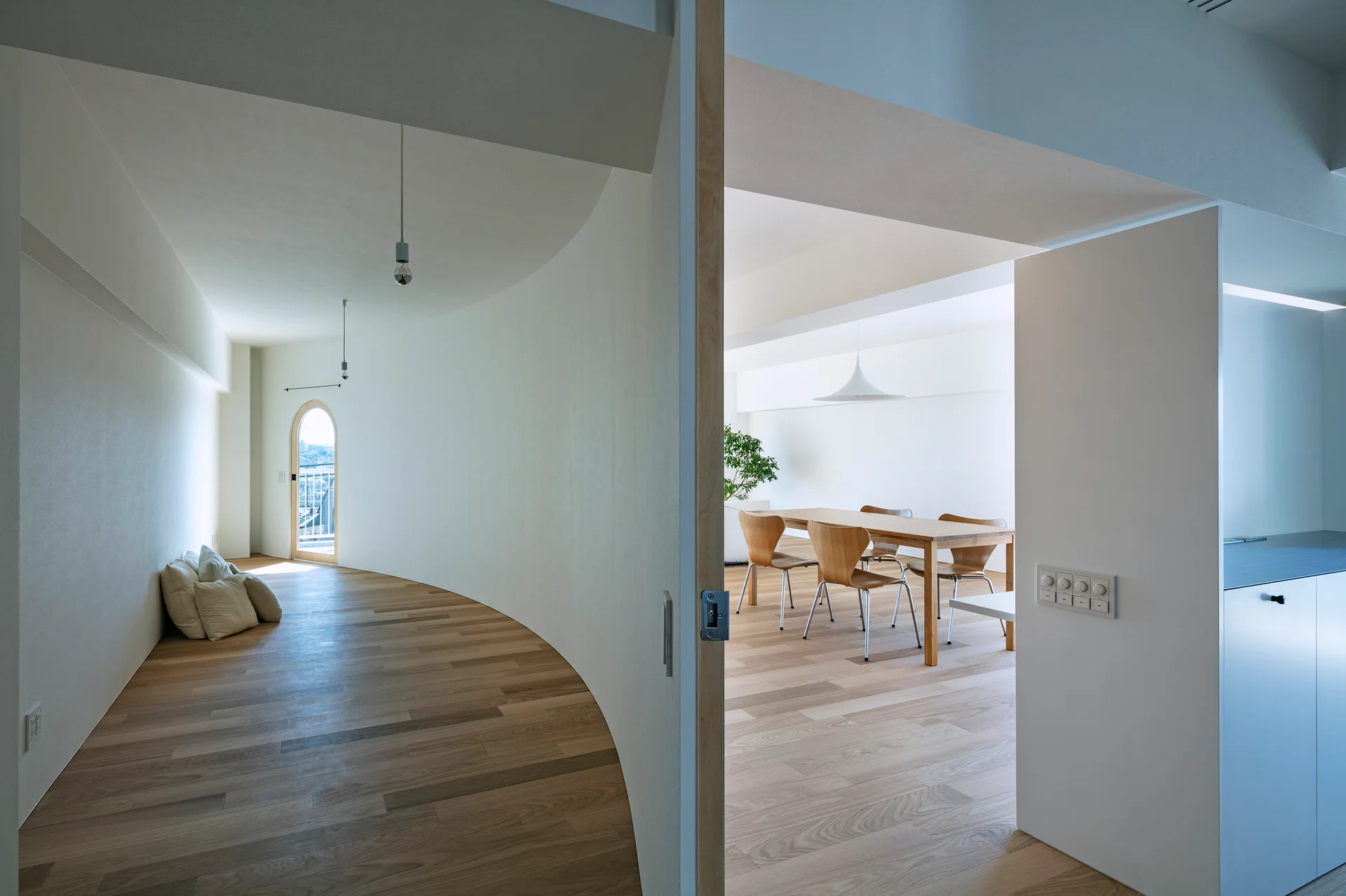 A bright minimalist dining area with a wooden table and chairs is visible past a large, curved white wall partition and light wood floors.