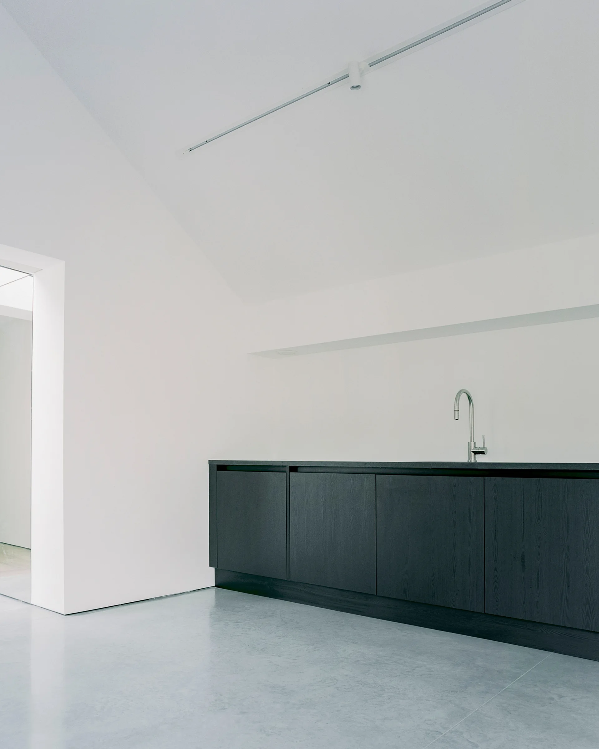 A minimalist kitchen displays dark wood cabinets with a metal faucet, white walls, and a grey concrete floor.