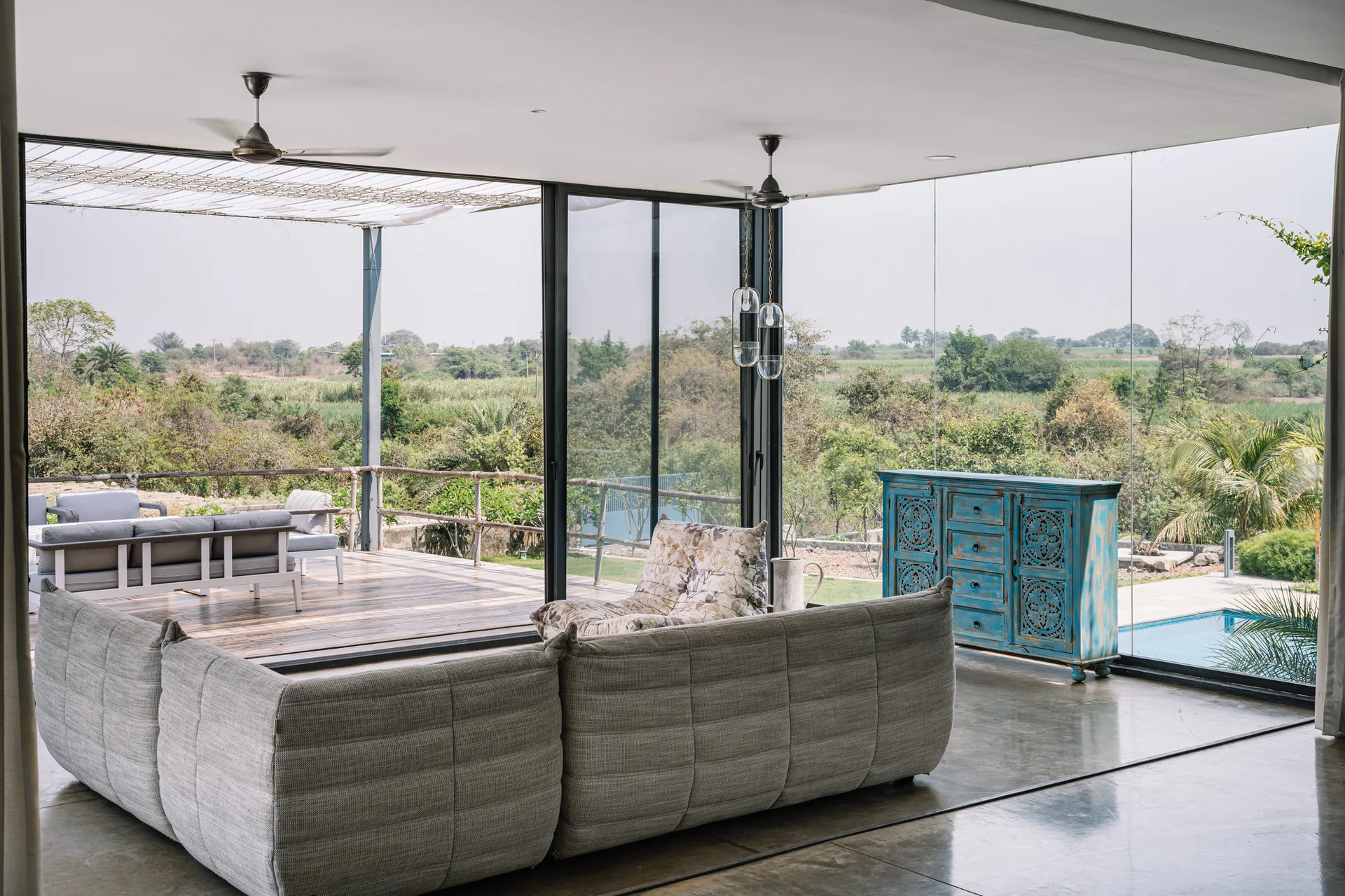 A modern living room features a textured grey modular sofa, a carved blue cabinet, and floor-to-ceiling glass walls opening onto a wooden deck.