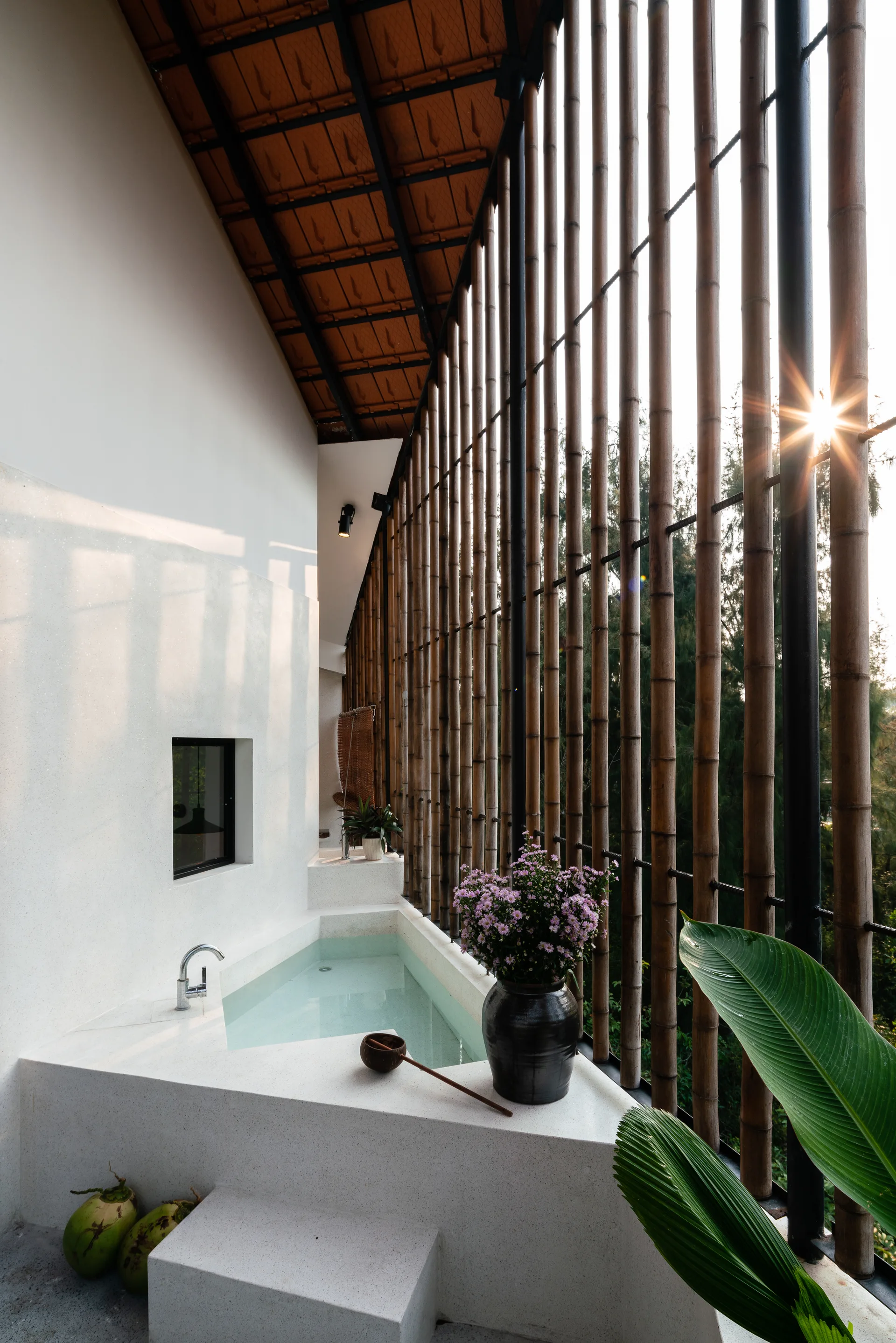 An open-air bathing area features a built-in terrazzo tub positioned next to a tall vertical bamboo screen wall with filtering sunlight.