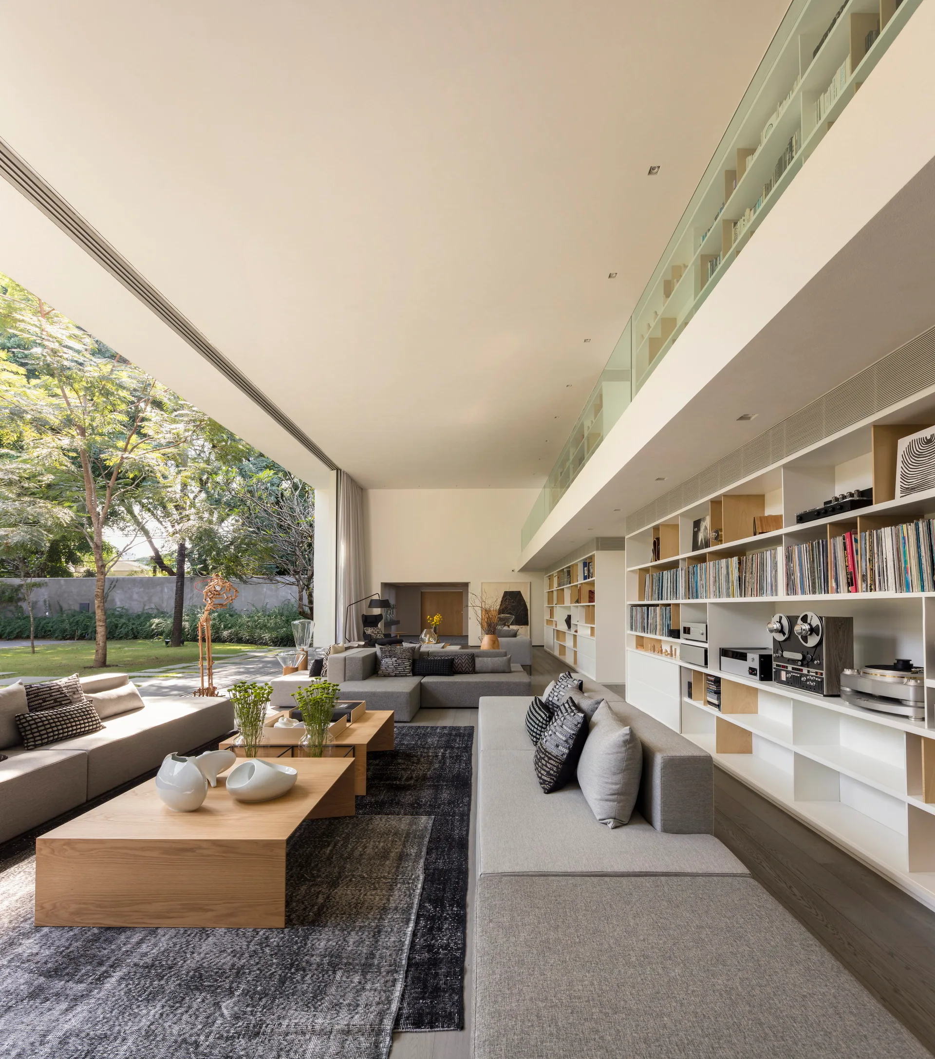 A modern living room features long grey sectional sofas, low wooden coffee tables, and a large built-in shelving wall opening to a grassy backyard.
