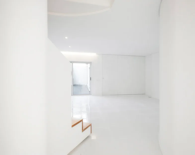 A minimalist white interior hallway features glossy white reflective floors, curved walls, and a staircase with wooden treads.