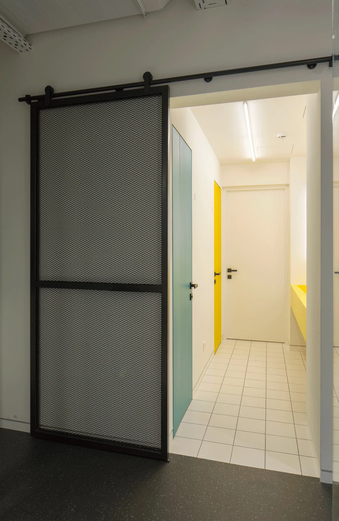 A narrow hallway with white square floor tiles features a sliding black mesh door and colorful interior doors in teal and yellow.