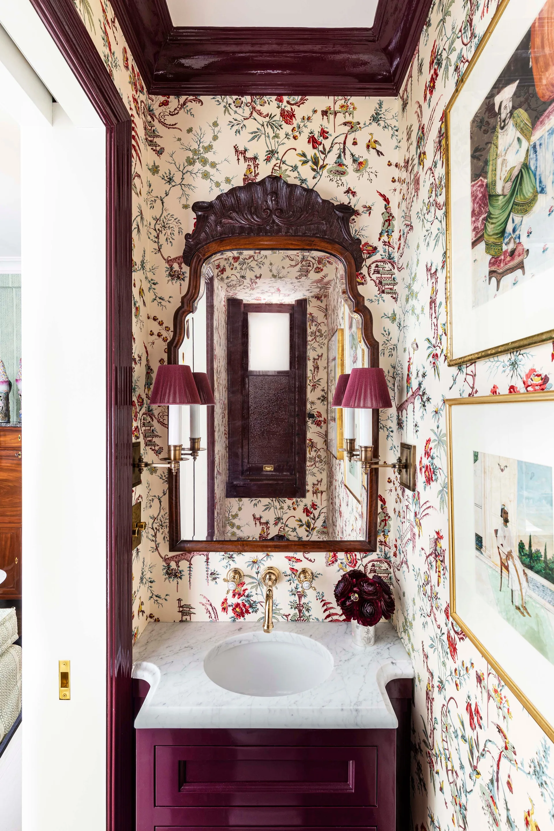A small powder room features Chinoiserie wallpaper, a marble-topped burgundy vanity, and an ornate dark wood mirror flanked by shaded sconces.
